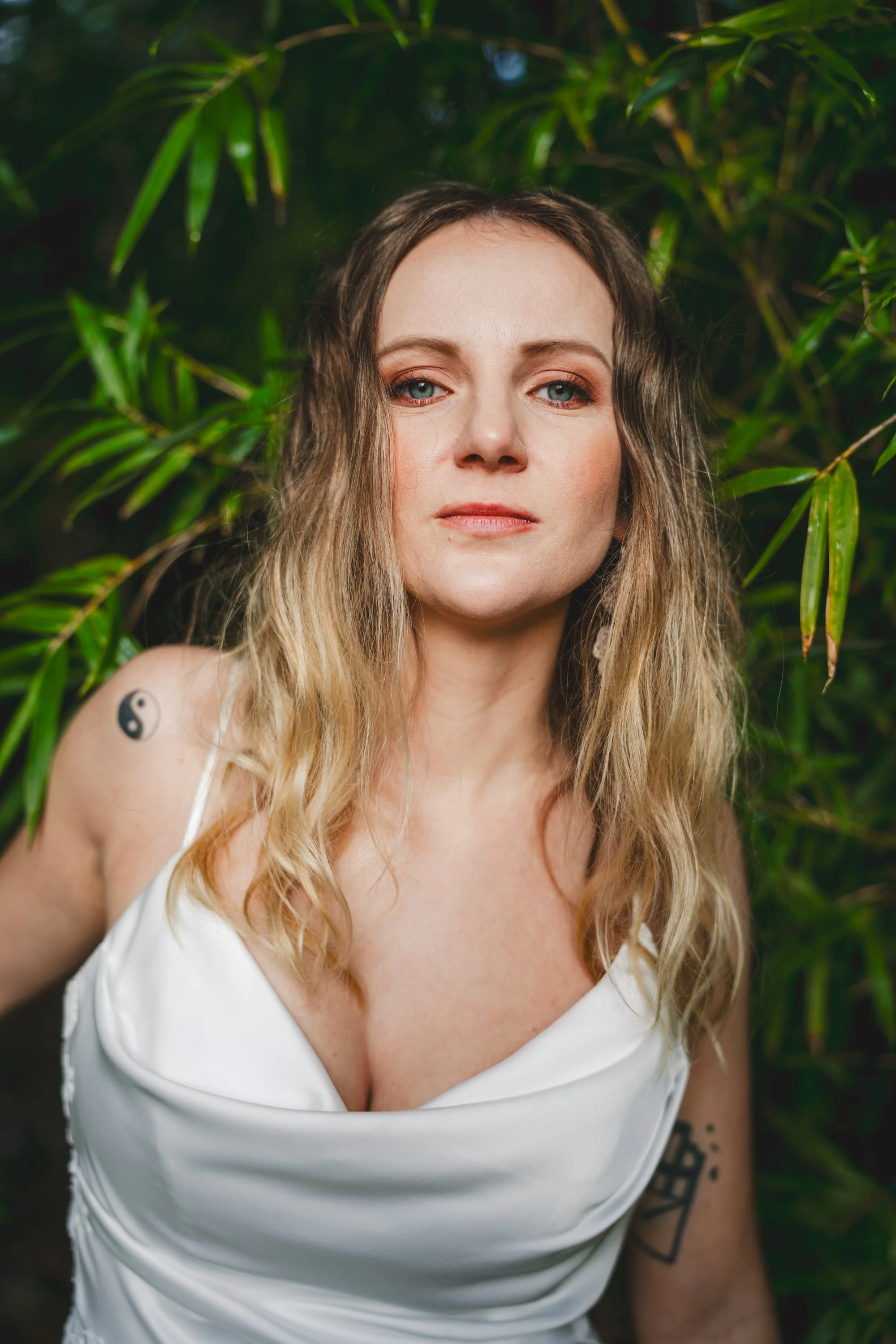 A woman with wavy blonde hair and tattoos on her shoulder and arm, wearing a white dress, standing in front of green bamboo leaves.