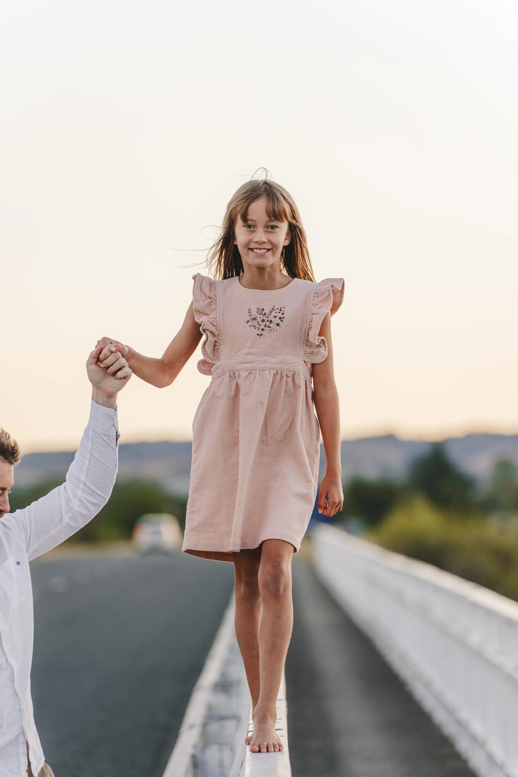 A young girl walking barefoot on a narrow ledge with a man holding her hand for support, outdoors during sunset.
