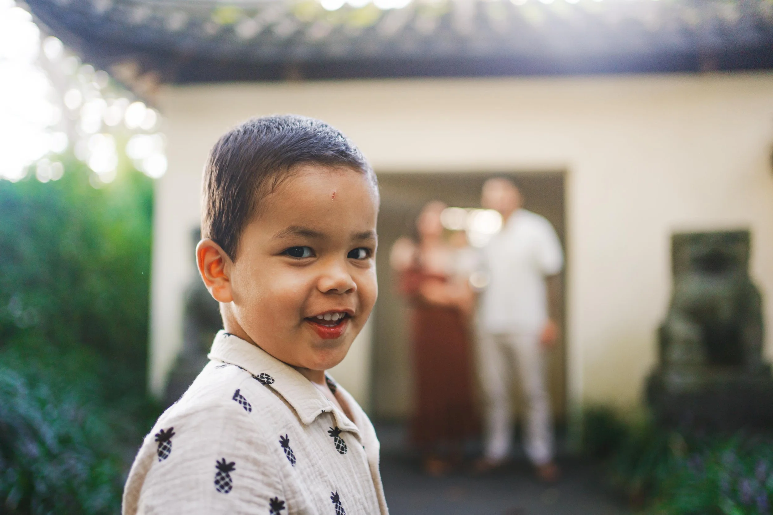 Smiling young boy with short dark hair wearing a cream shirt with pineapple illustrations, standing outdoors with a blurred background showing a house and three people.