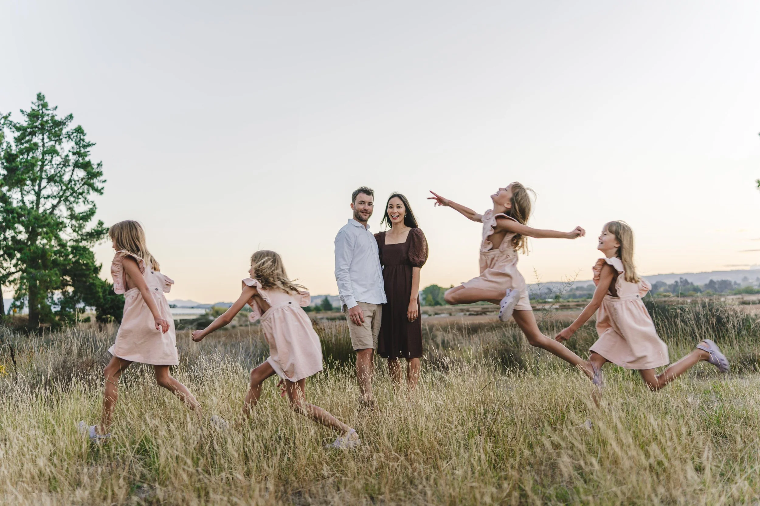 Family of seven, including two adults and five young girls, playing and jumping in a grassy field during sunset.