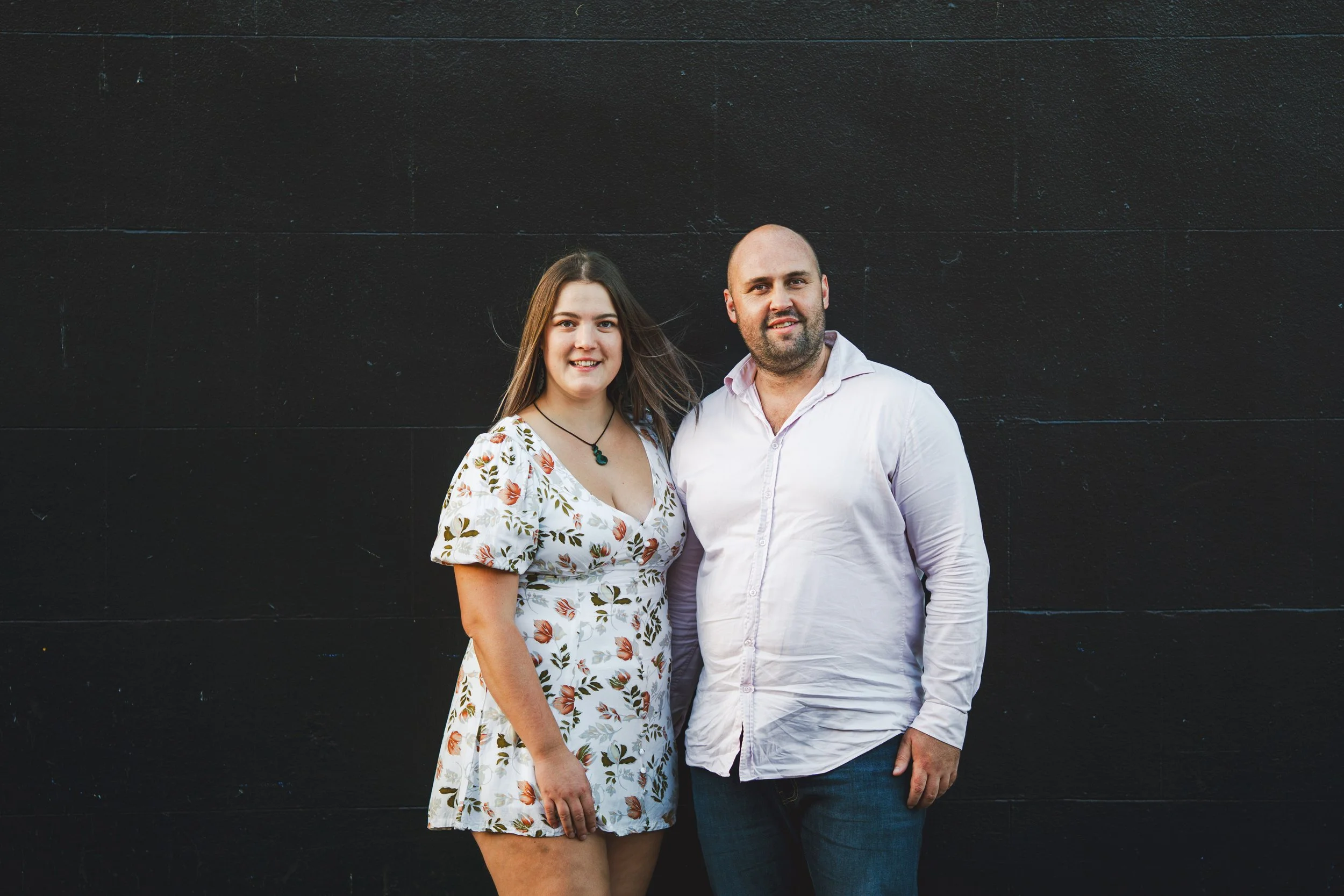 A young woman and a man standing together in front of a black wall, smiling at the camera.