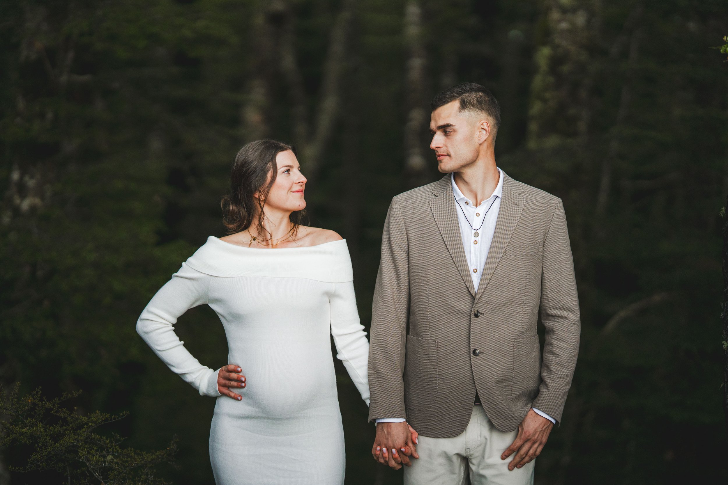 A woman in a white off-shoulder dress and a man in a gray blazer and white pants holding hands outdoors in a wooded area.