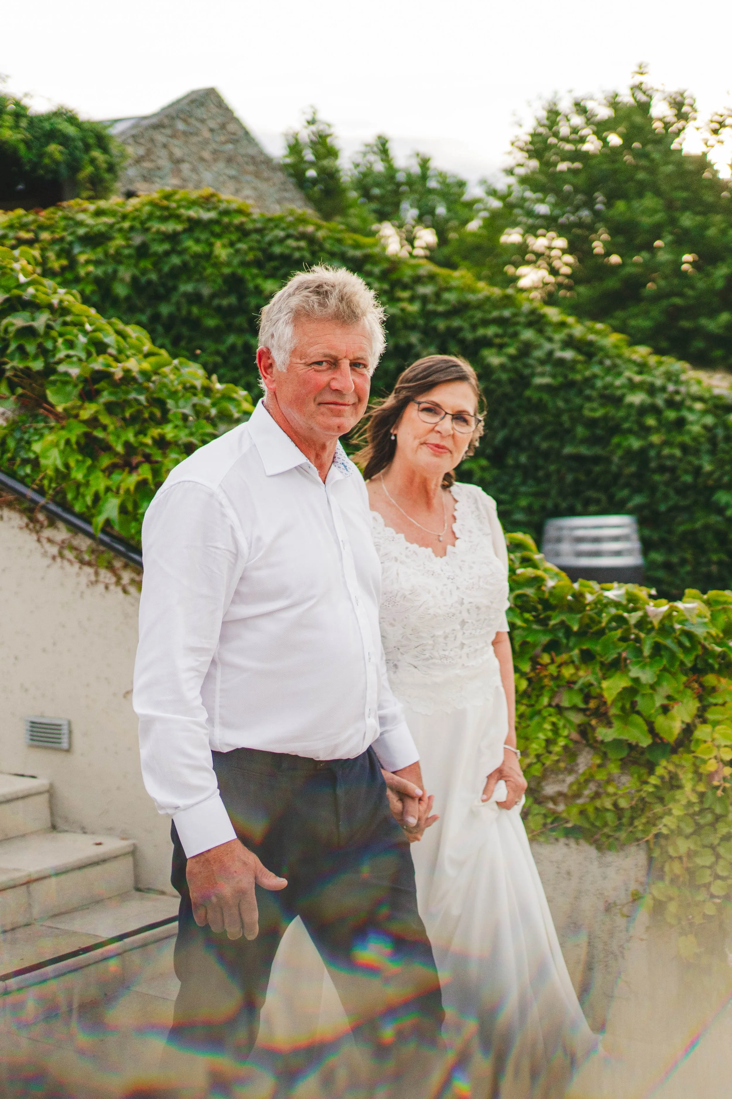 A mature couple walking hand-in-hand outdoors, with greenery and a stone building in the background.
