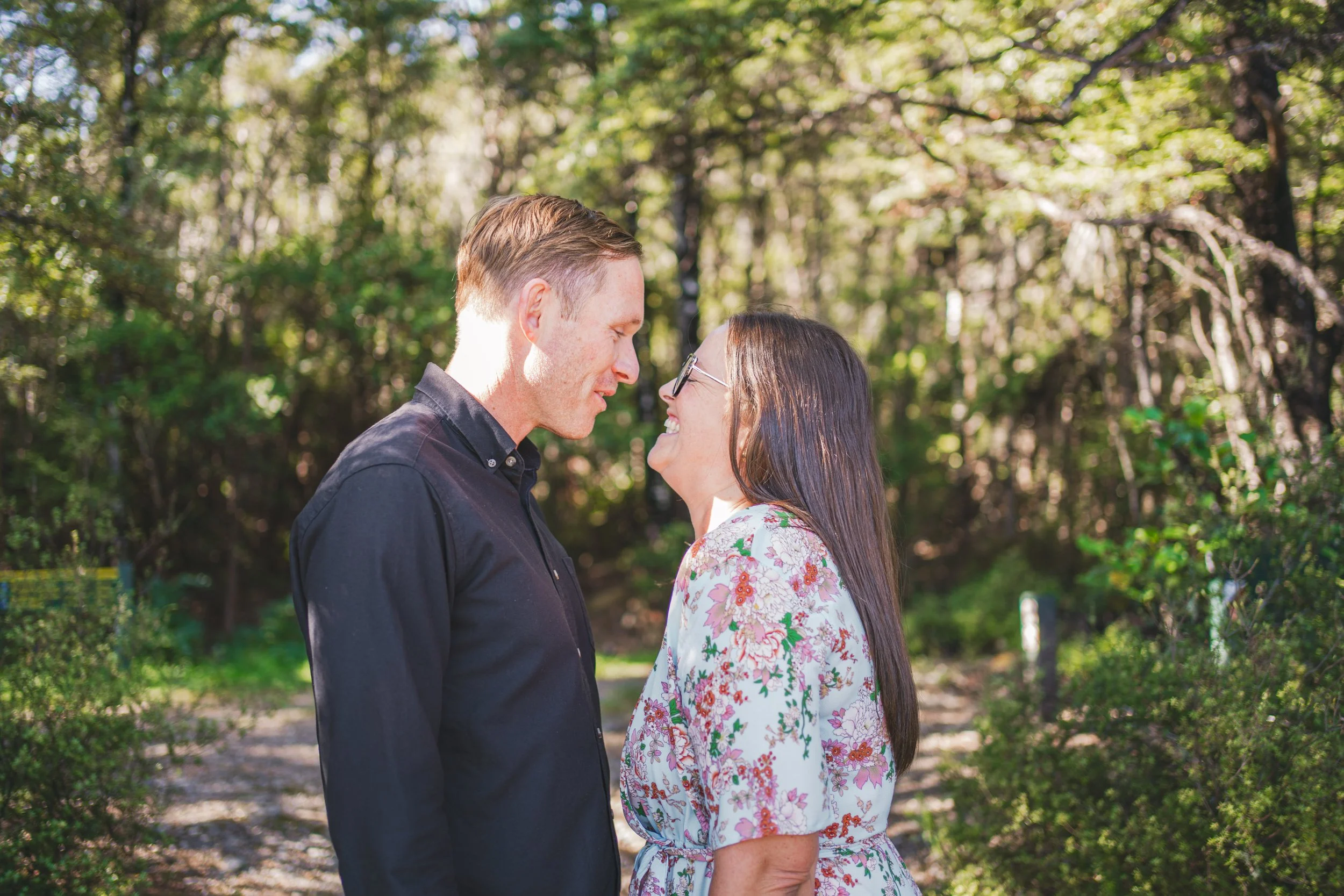 A couple standing close together outdoors in a wooded area, smiling and looking at each other.