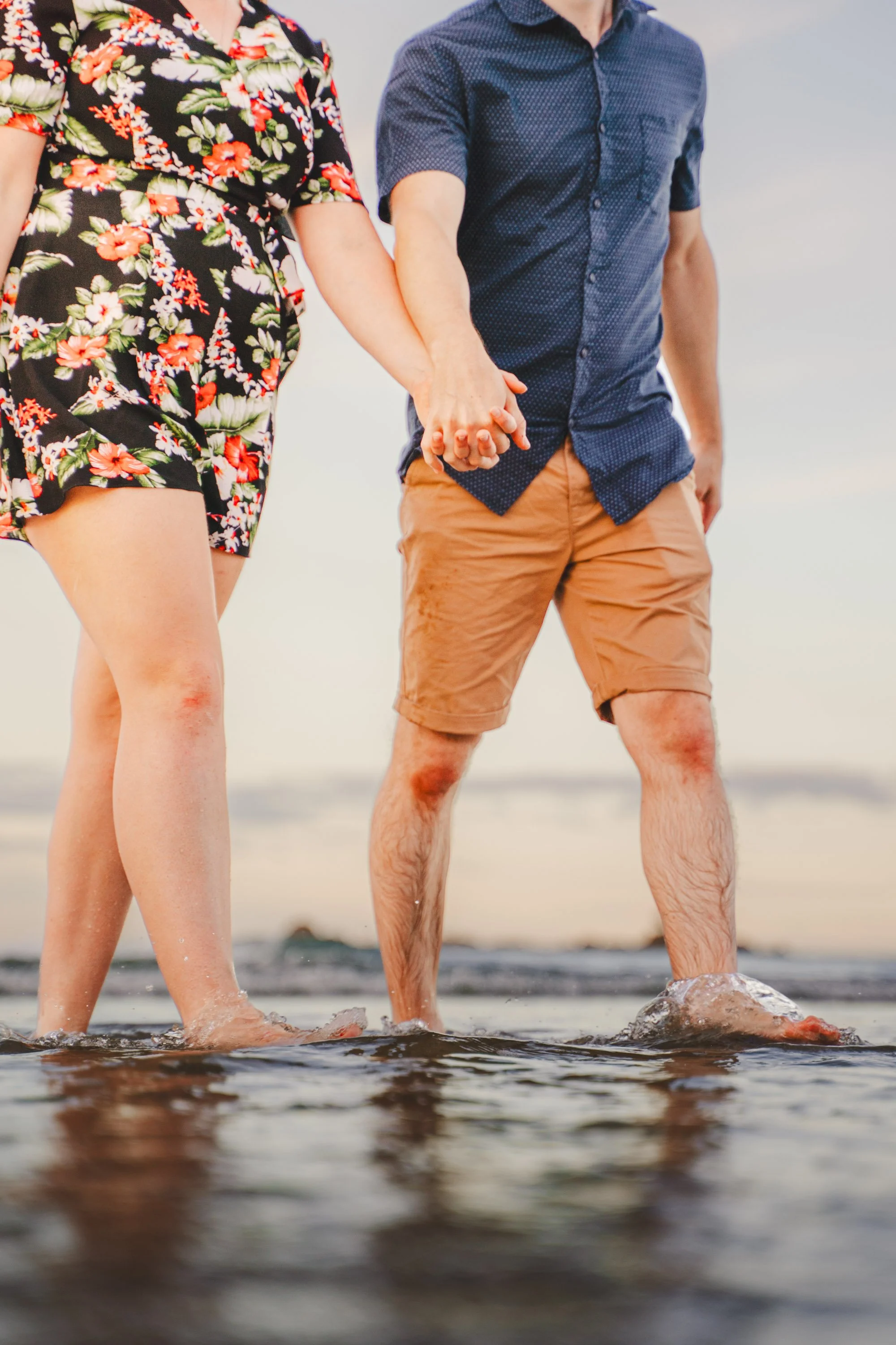 A couple holding hands and standing in shallow water at the beach during sunset, with only their lower bodies visible.
