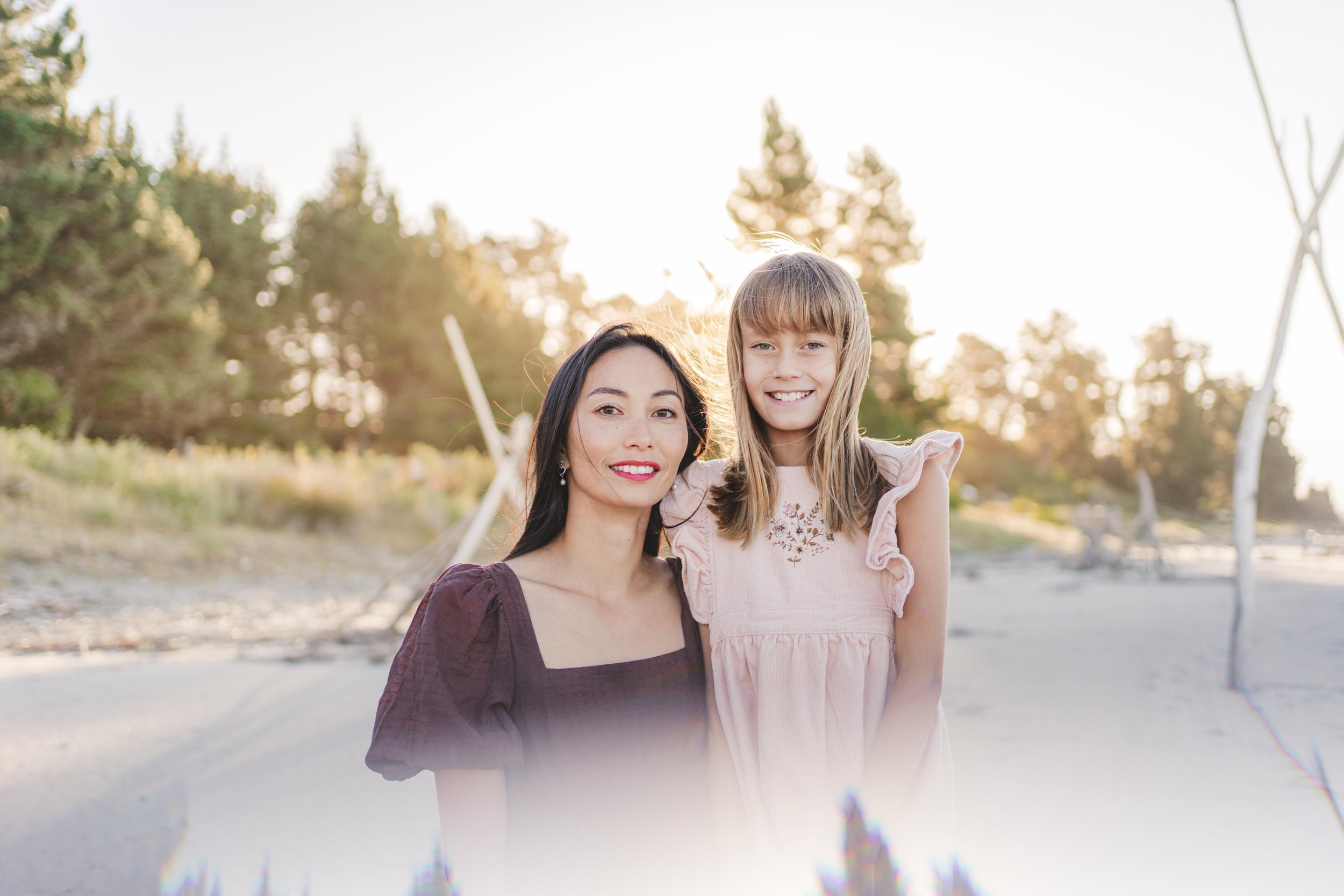 A woman and a young girl smiling on a beach at sunset, standing close together with trees and driftwood in the background.