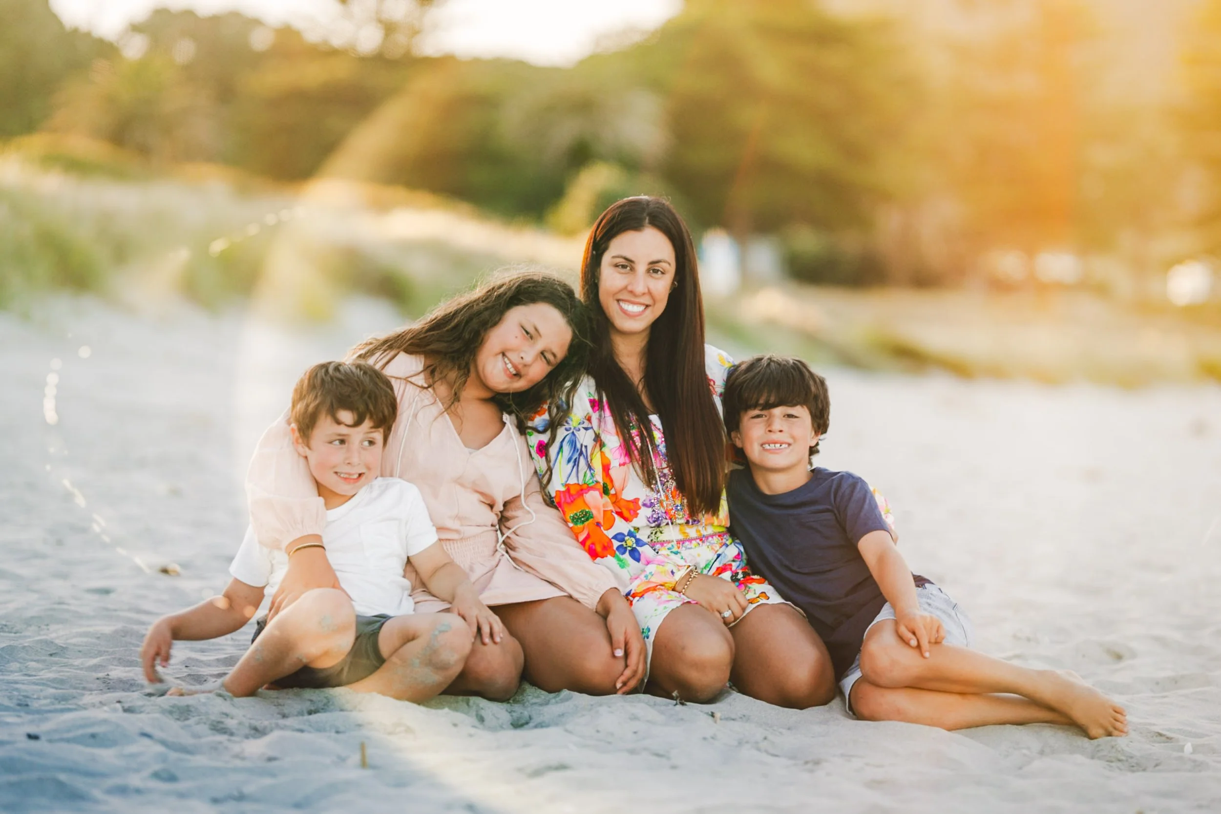 A woman and three children sitting together on a sandy beach with trees in the background during sunset, smiling.