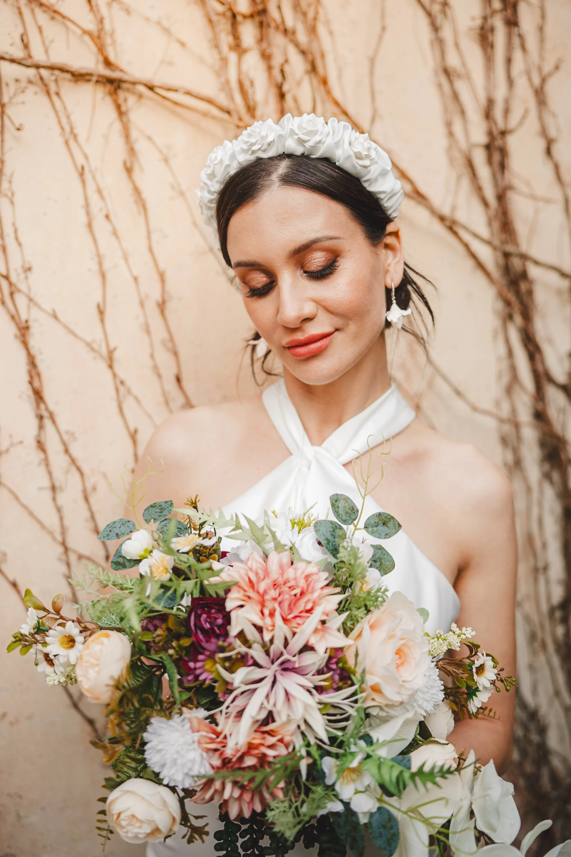 Bride with a floral headband holding a large bouquet of mixed flowers, standing in front of a wall with dried vines.