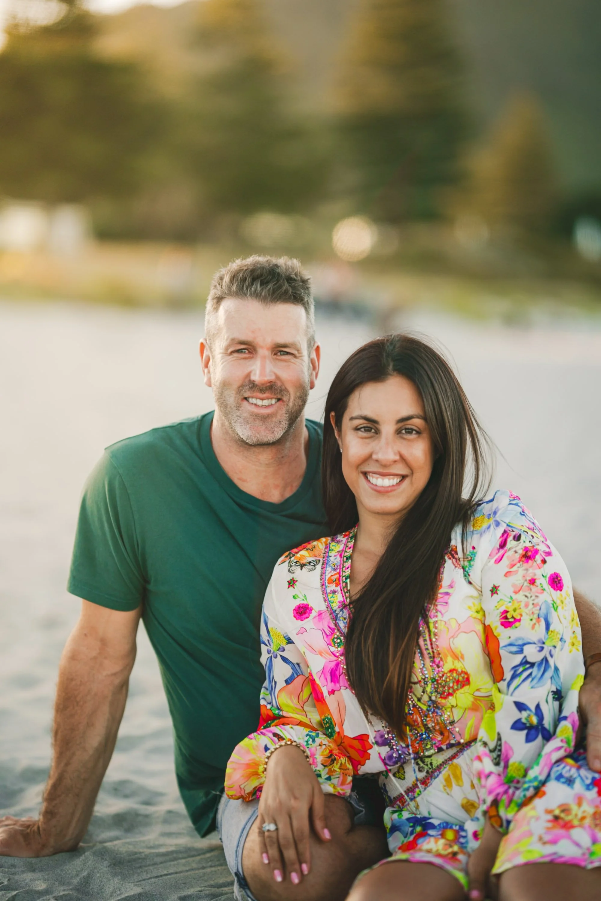 A smiling man and woman sitting on a sandy beach with trees and water in the background, during sunset.