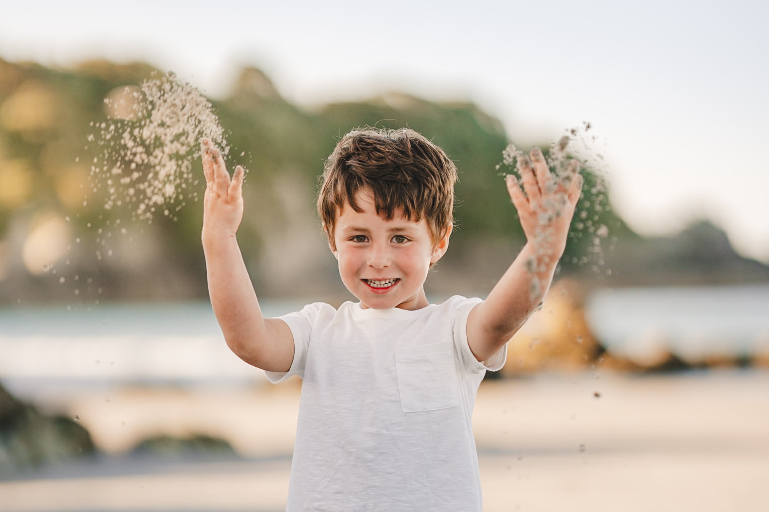 Child playing with sand at the beach, smiling and waving sand in the air.