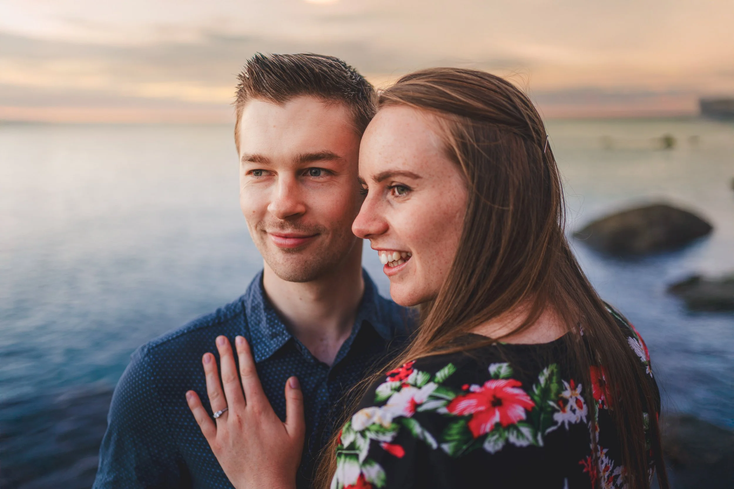 A couple standing by the water during sunset, with the woman showing off a wedding ring.
