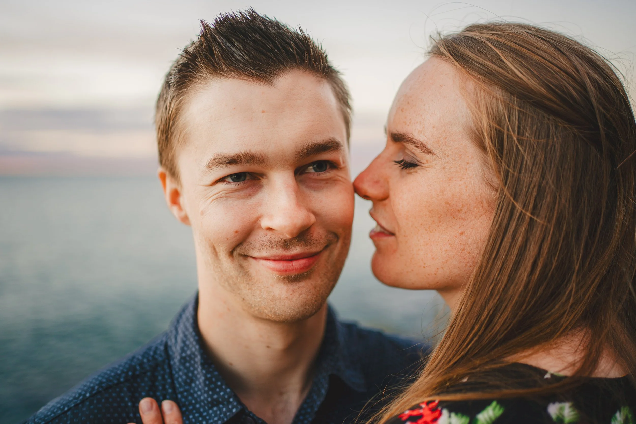 A smiling man and a woman with closed eyes are close together near a body of water during sunset.