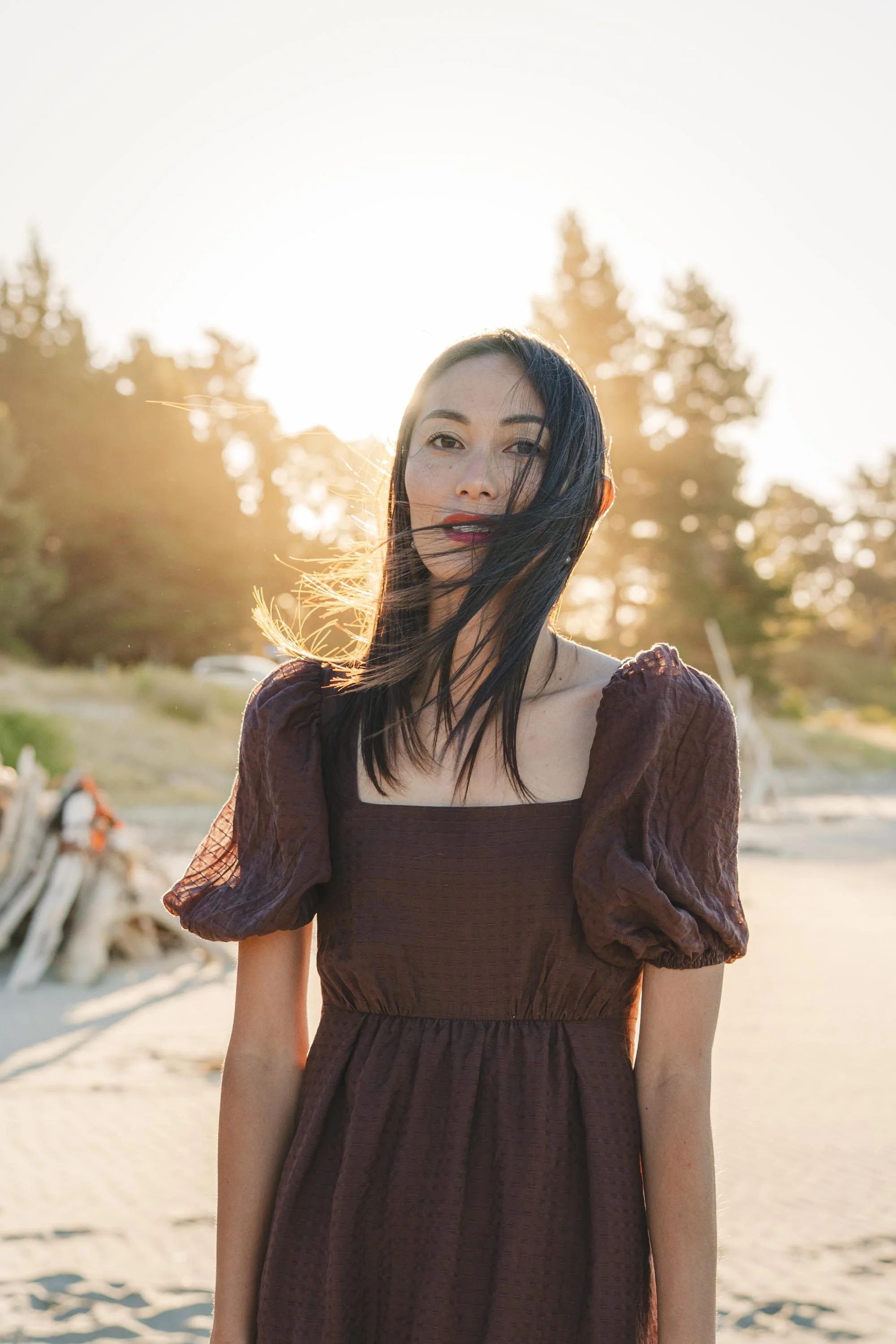 A woman with long dark hair and fair skin stands outdoors during sunset, wearing a dark purple dress with puffed sleeves, with trees and a sandy area in the background.
