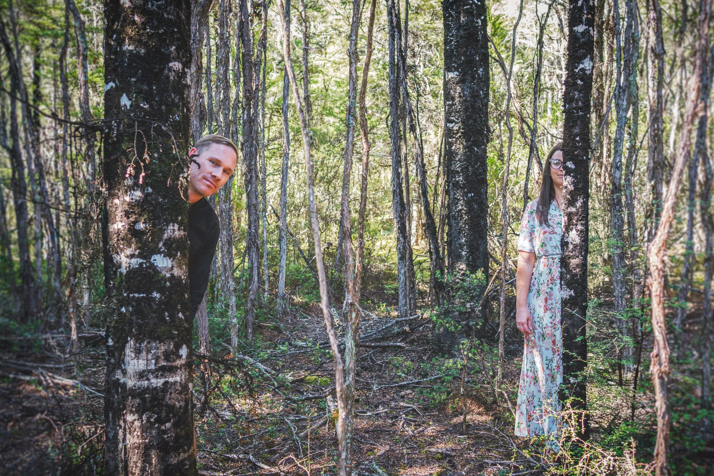 A man and a woman peeking from behind trees in a dense forest with green foliage and sunlight filtering through.