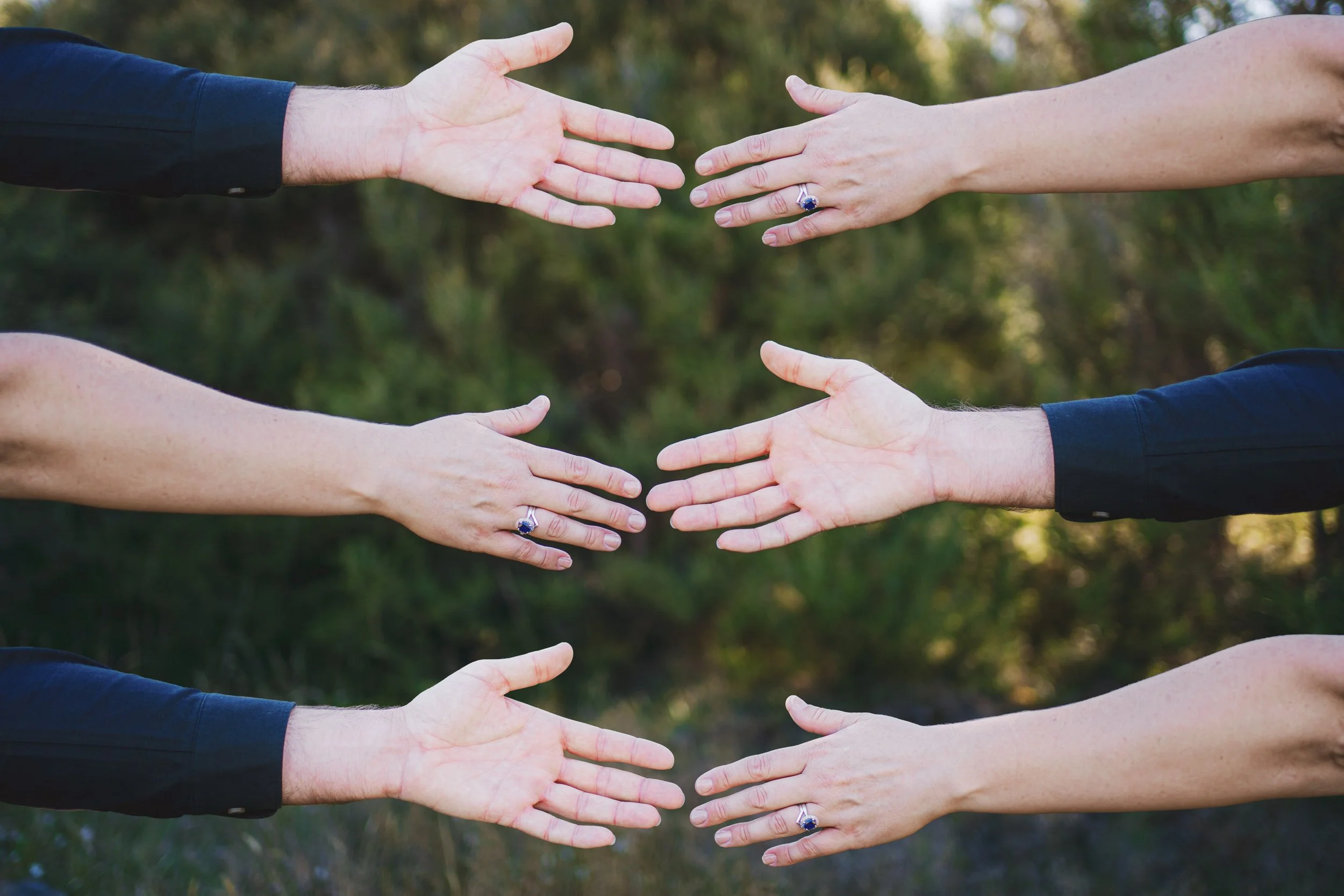Six hands reaching towards each other outdoors, three on the top and three on the bottom, with a blurred green natural background.