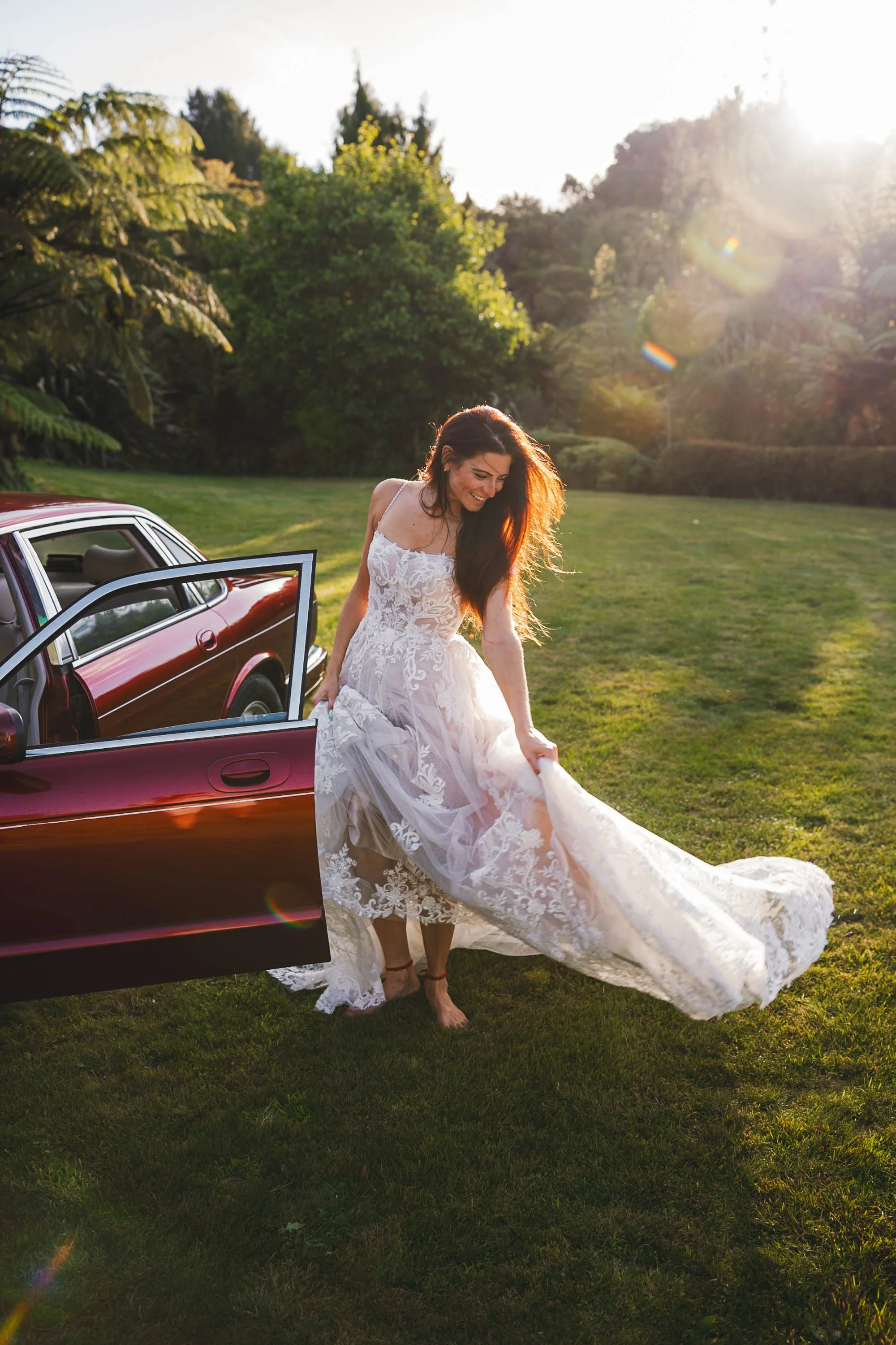 A woman in a wedding dress stepping out of a red car into a grassy field at sunset, smiling and holding her dress up as the sunlight creates a warm glow.