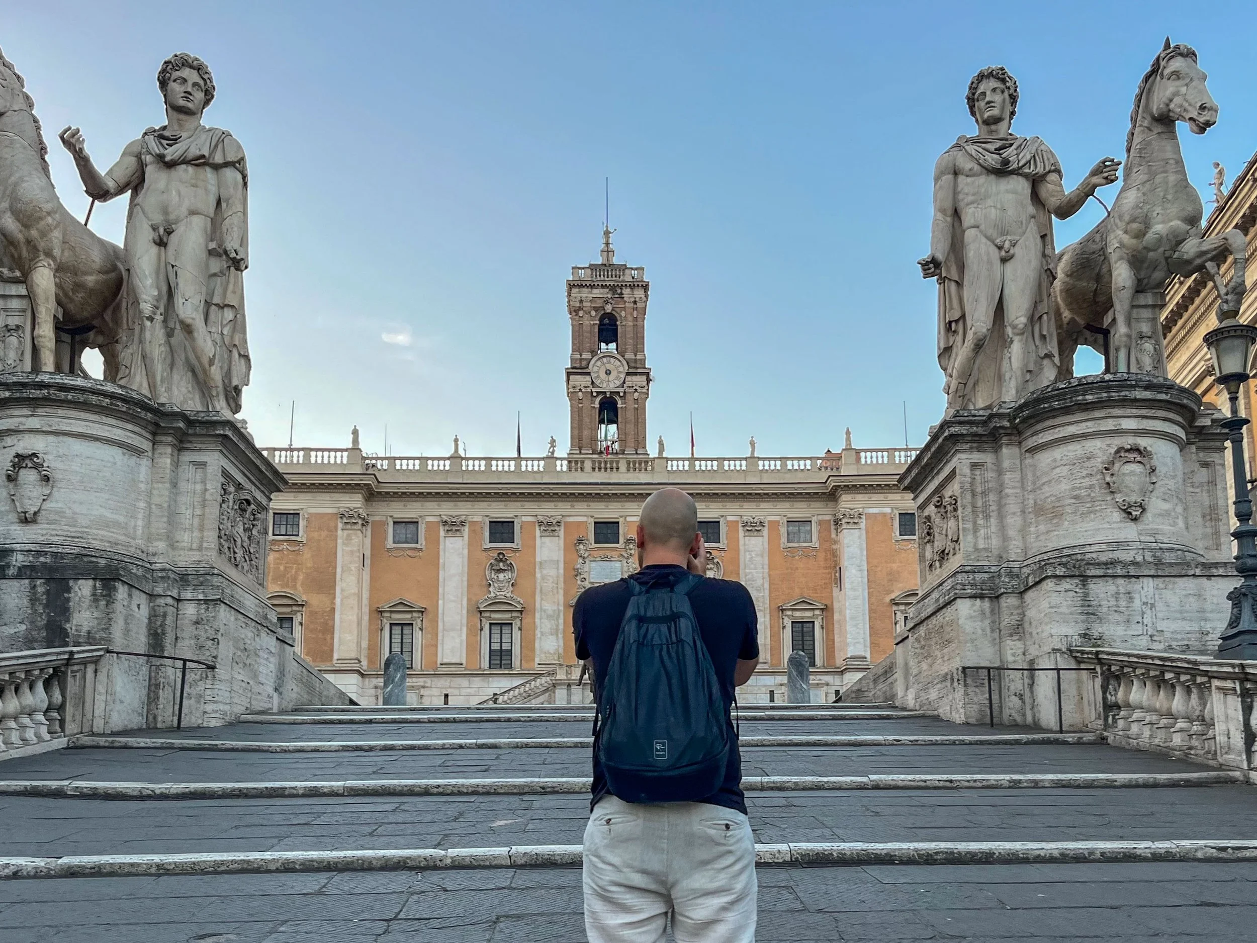 A man with a backpack taking a photograph in front of an ornate historic building with statues flanking the stairs.