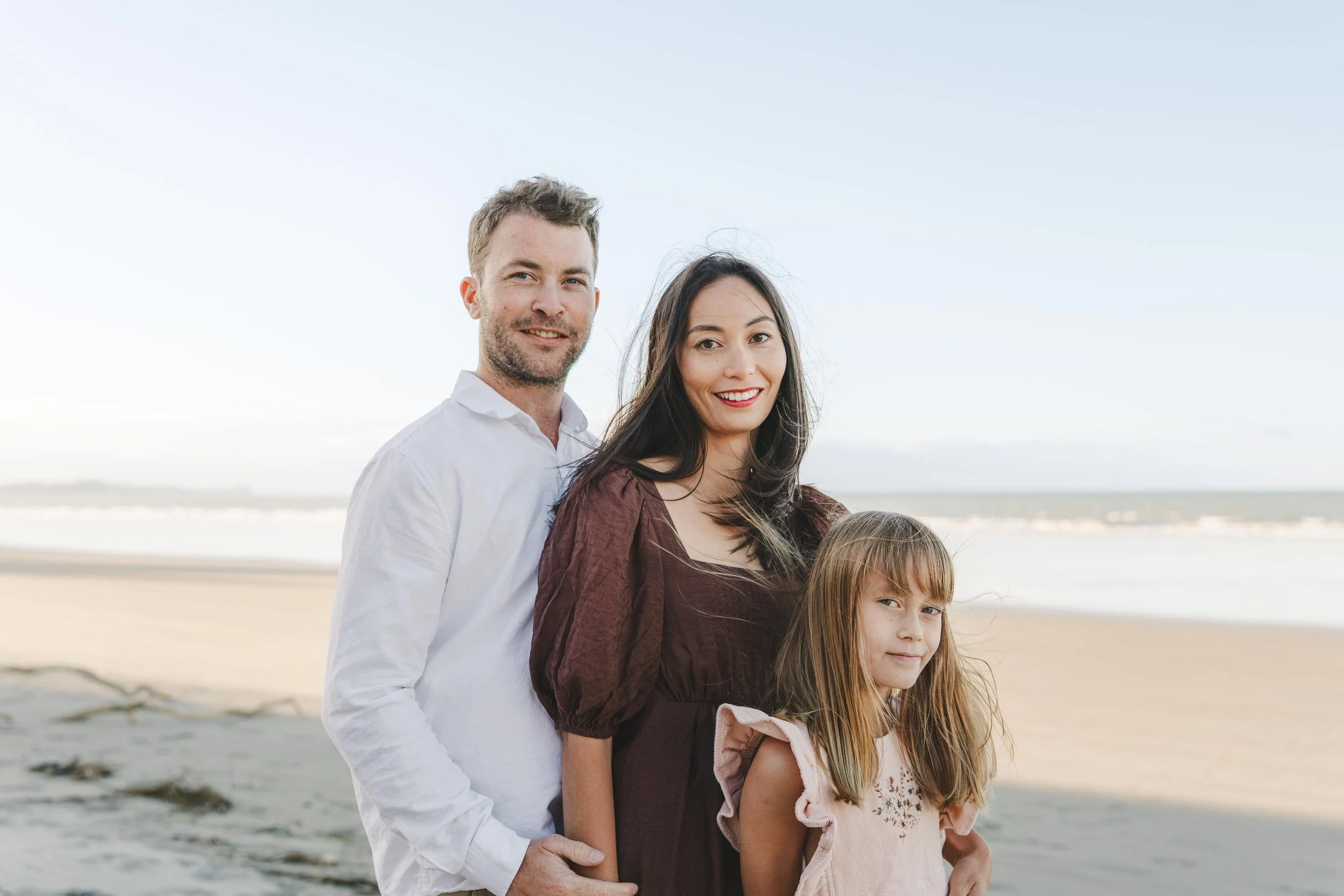 A family of three standing on the beach, with the ocean and sky in the background, smiling at the camera.
