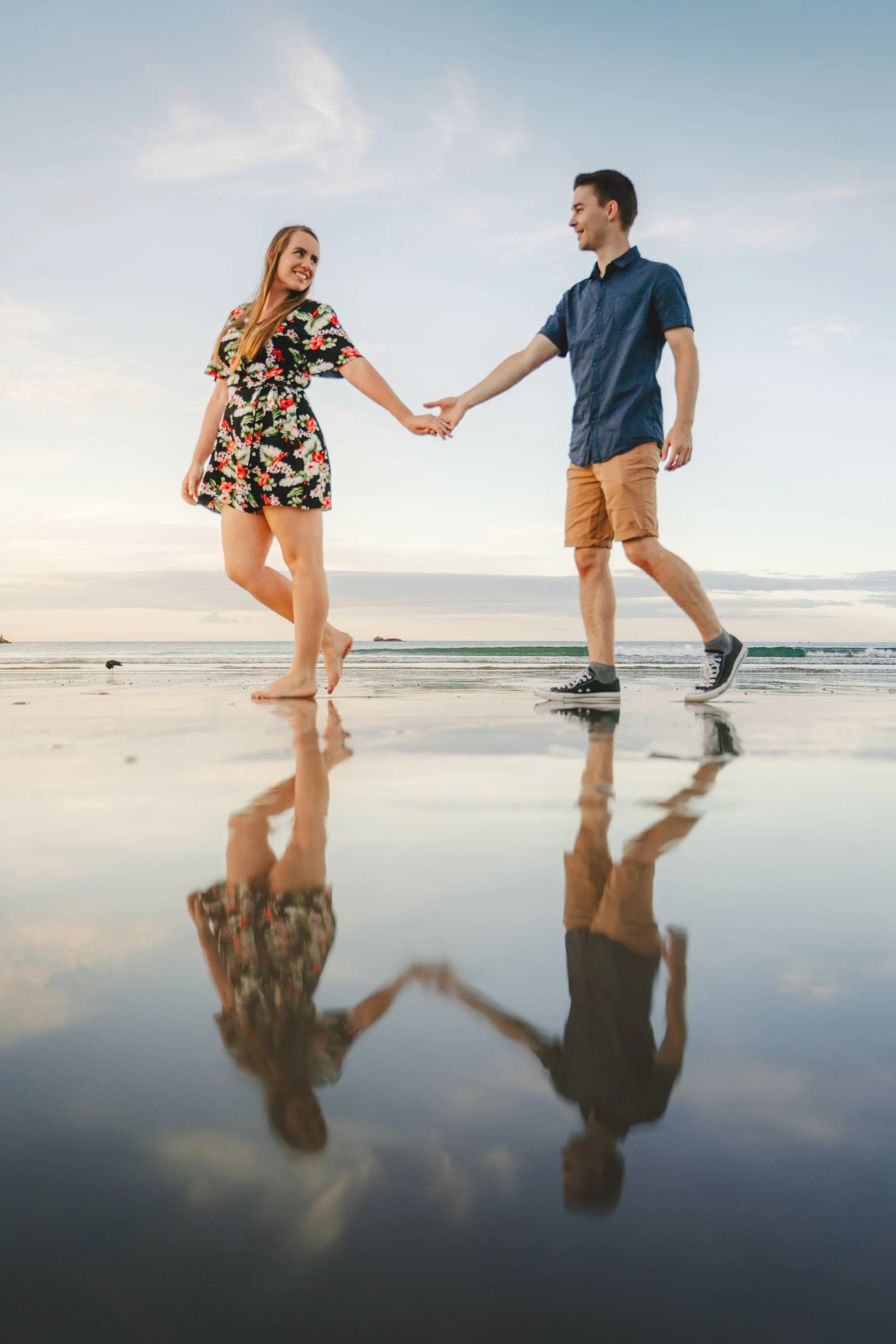 A young couple holding hands and dancing on a beach during sunset, with their reflection visible on the wet sand.