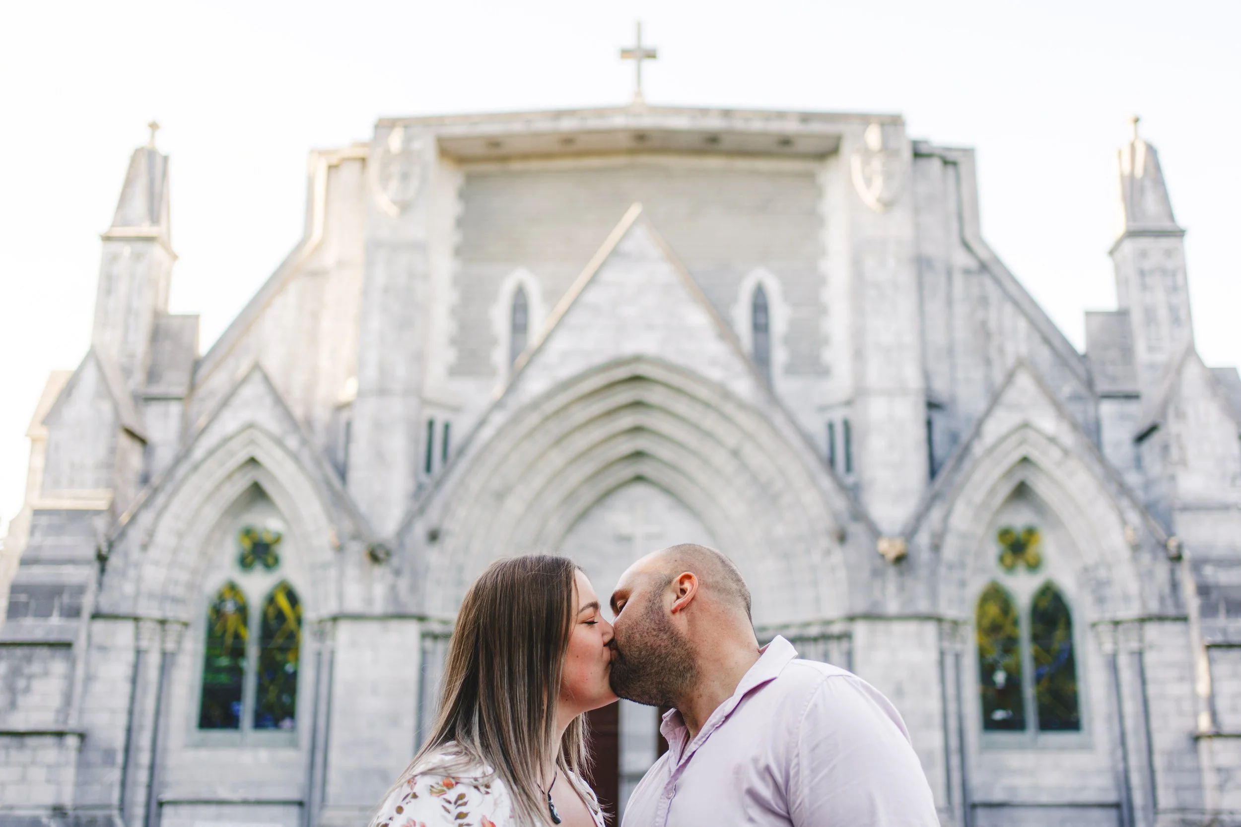 A couple kissing in front of a historic church with gothic architecture and stained glass windows.