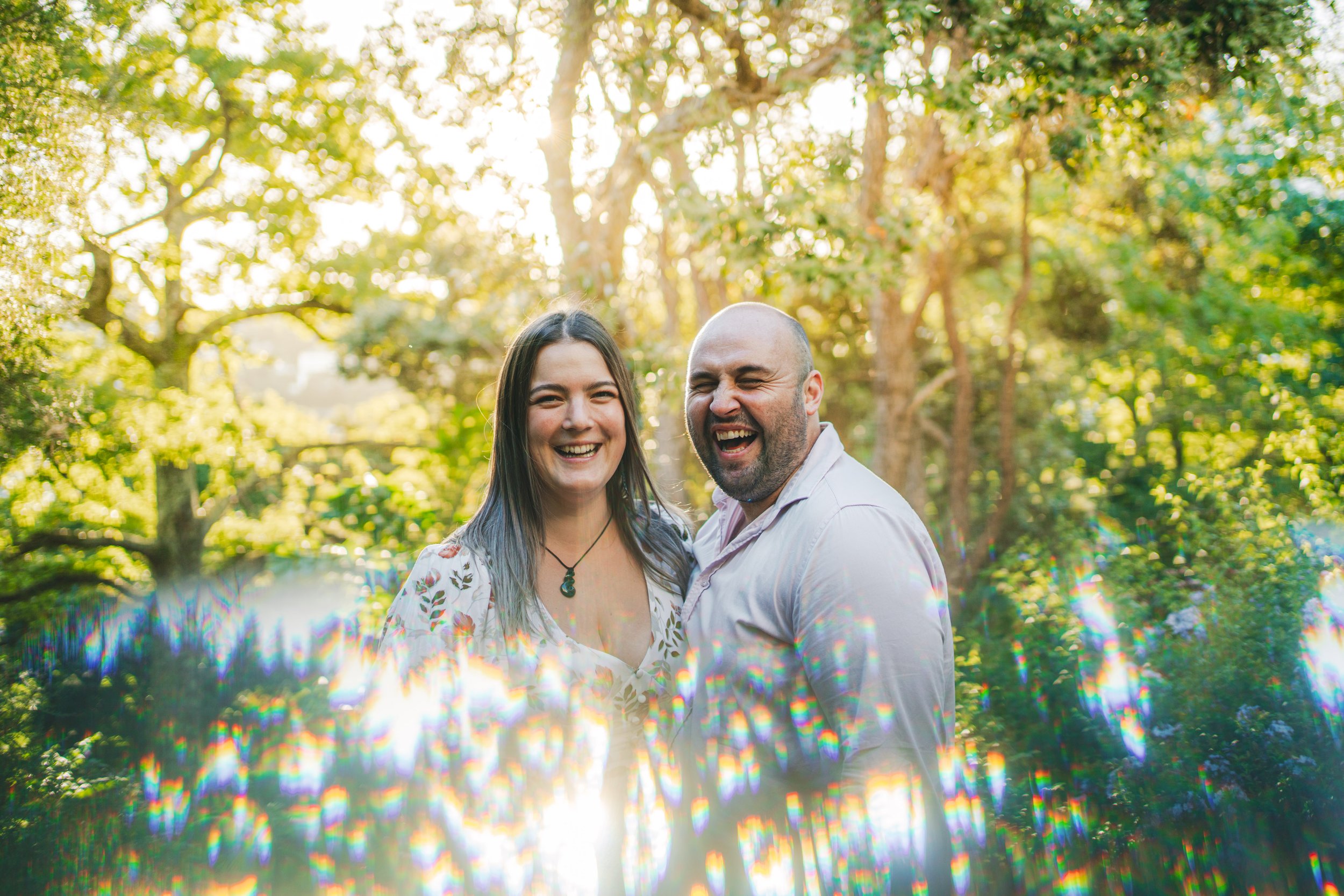 A smiling woman and a laughing man standing close together outdoors in a sunny, green, wooded area.