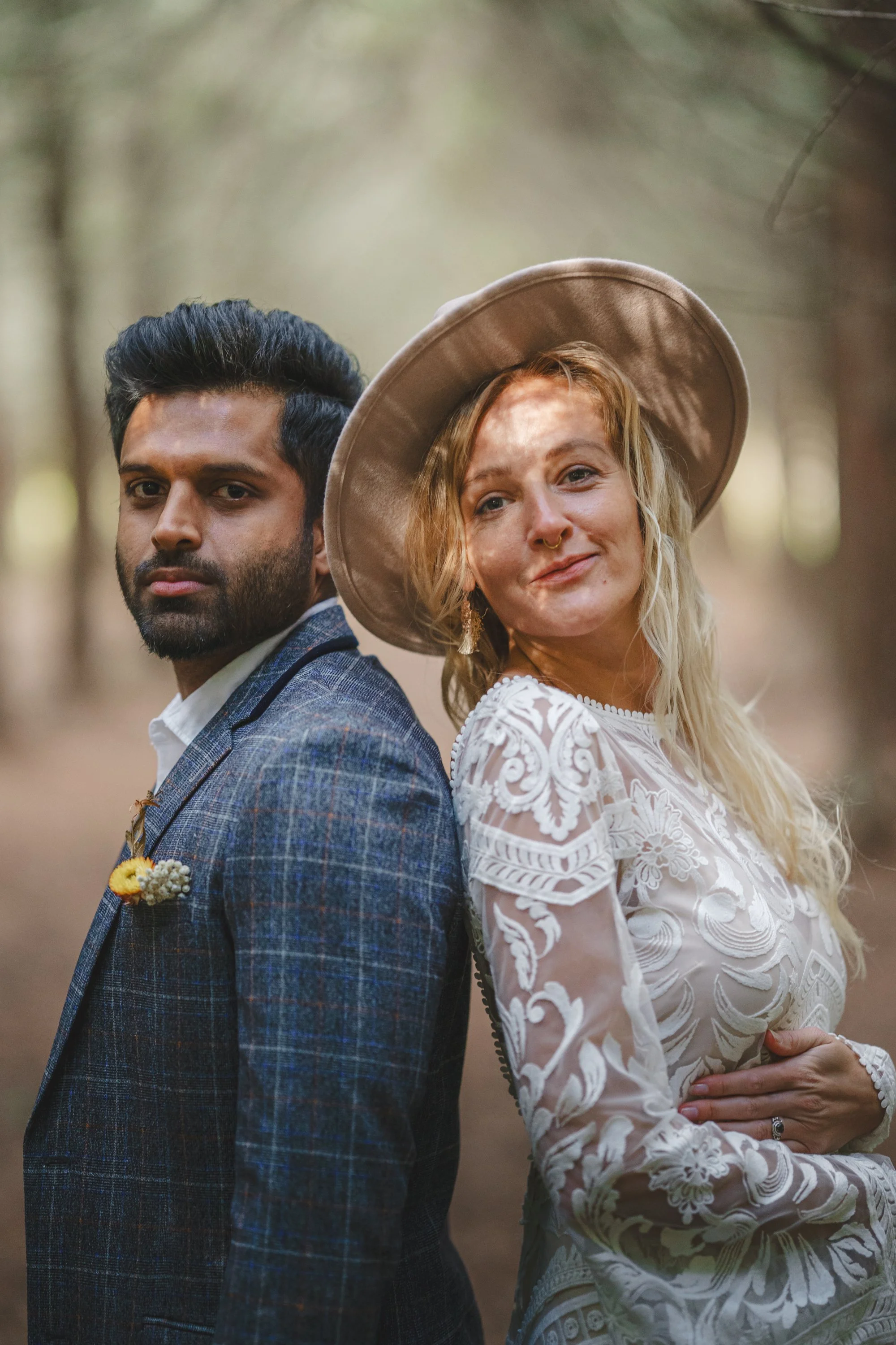 A man and woman standing back to back in a forest. The man has dark hair and a beard, dressed in a plaid suit. The woman has blonde hair, a wide-brimmed hat, and wears a white embroidered dress. She has a septum piercing and a flower ring on her fing