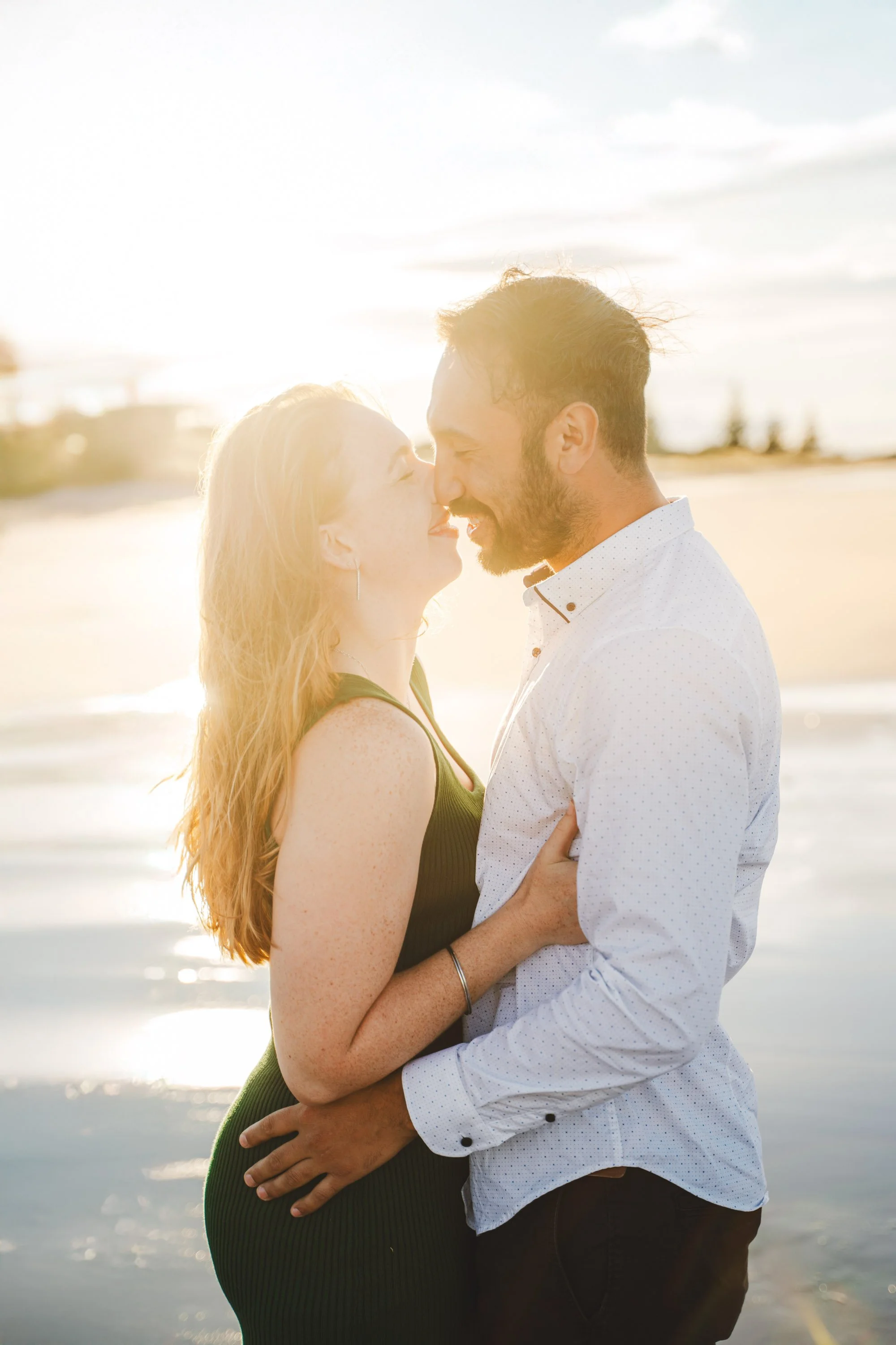 A couple is standing close together on a beach during sunset, about to kiss, with ocean and sky in the background.