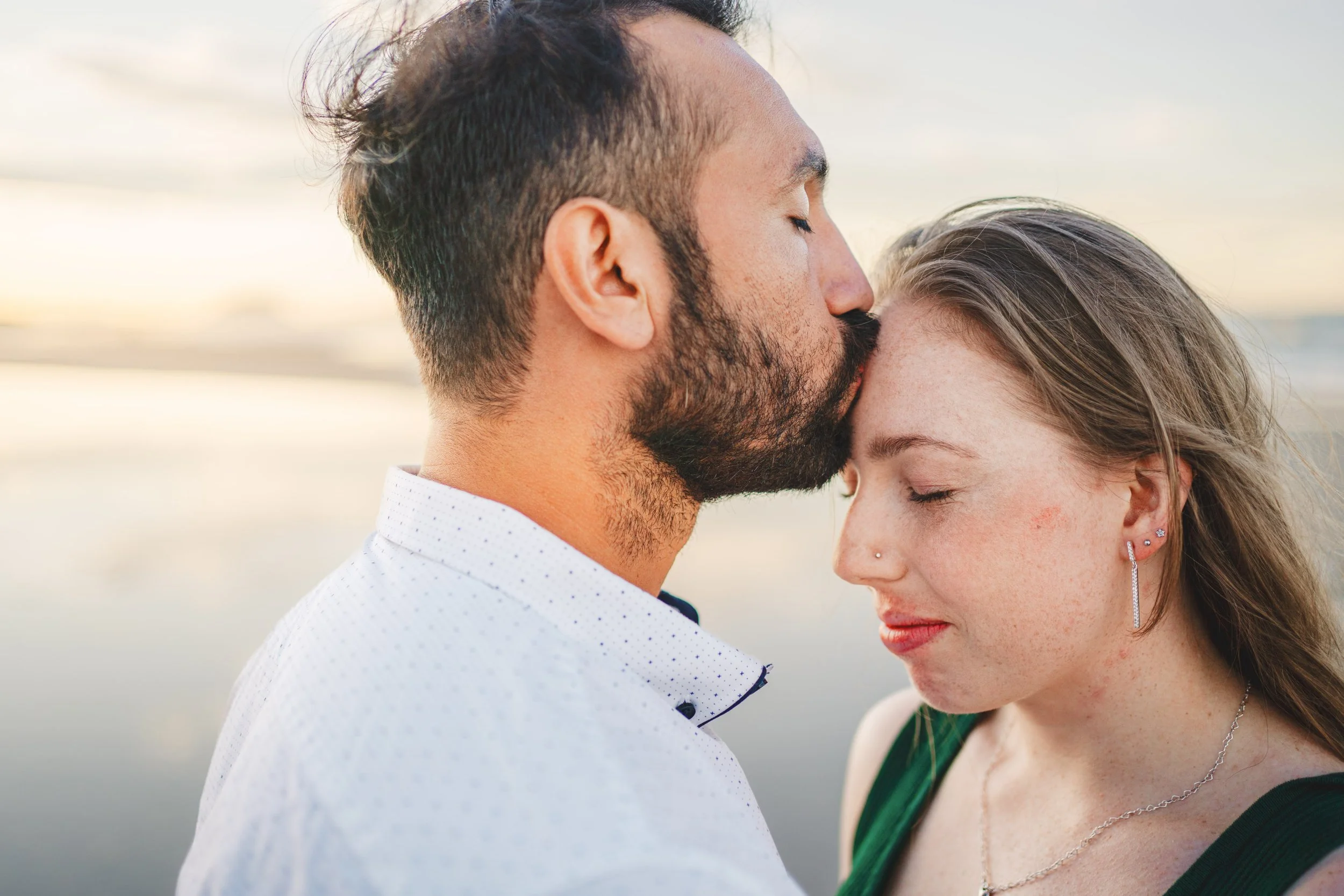 A man is kissing a woman on her forehead near a body of water at sunset, both with their eyes closed in a tender moment.