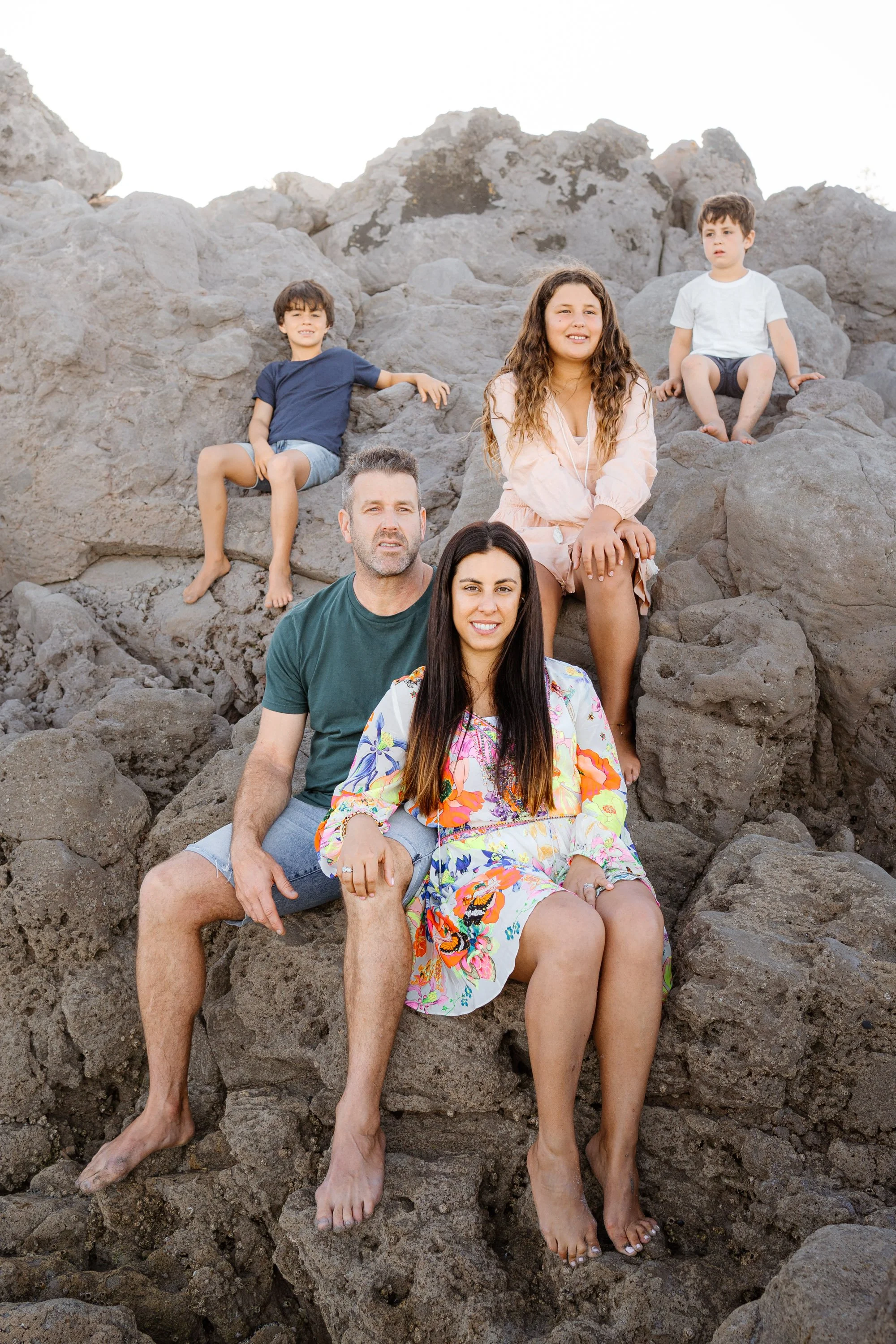 A family of five, including two adult women and three young boys, sitting on large rocks outdoors during daytime. They are barefoot and dressed in casual, summery clothing.