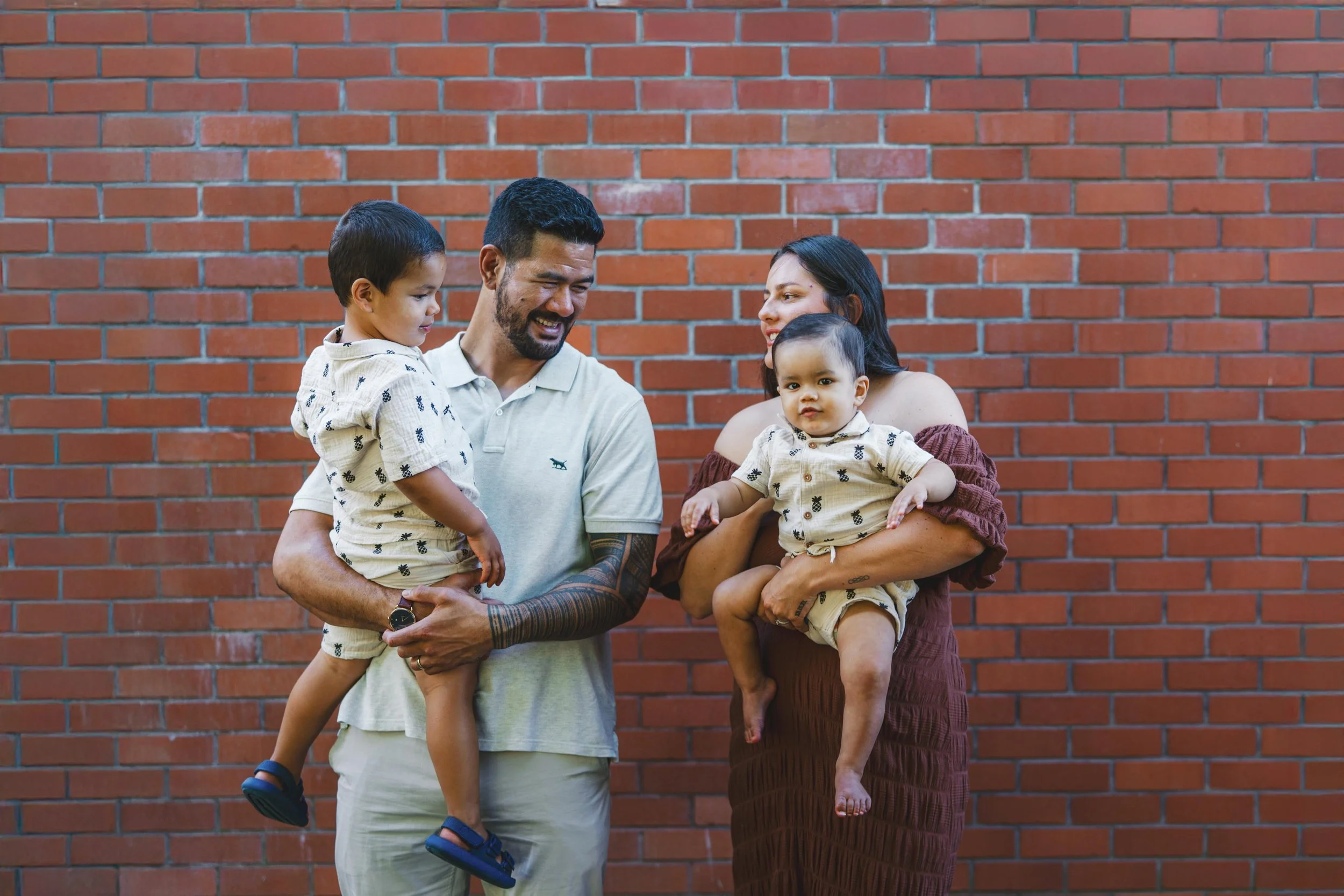 A family of four standing against a brick wall, smiling and interacting with each other. The father is holding a young boy, and the mother is holding a toddler girl. All appear to be happy and enjoying a moment together.