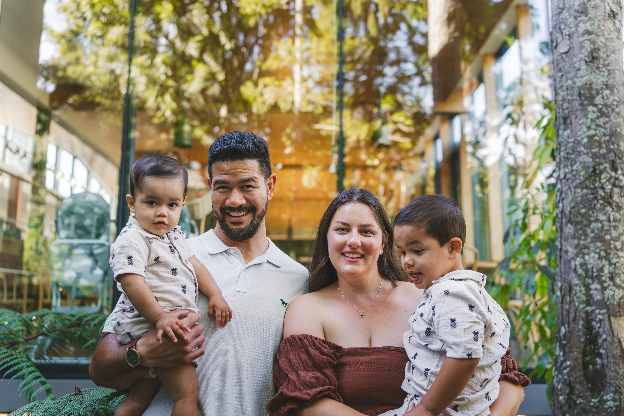 A smiling family of four posing outdoors in a lush garden, holding two young children, with trees and a wooden structure in the background.