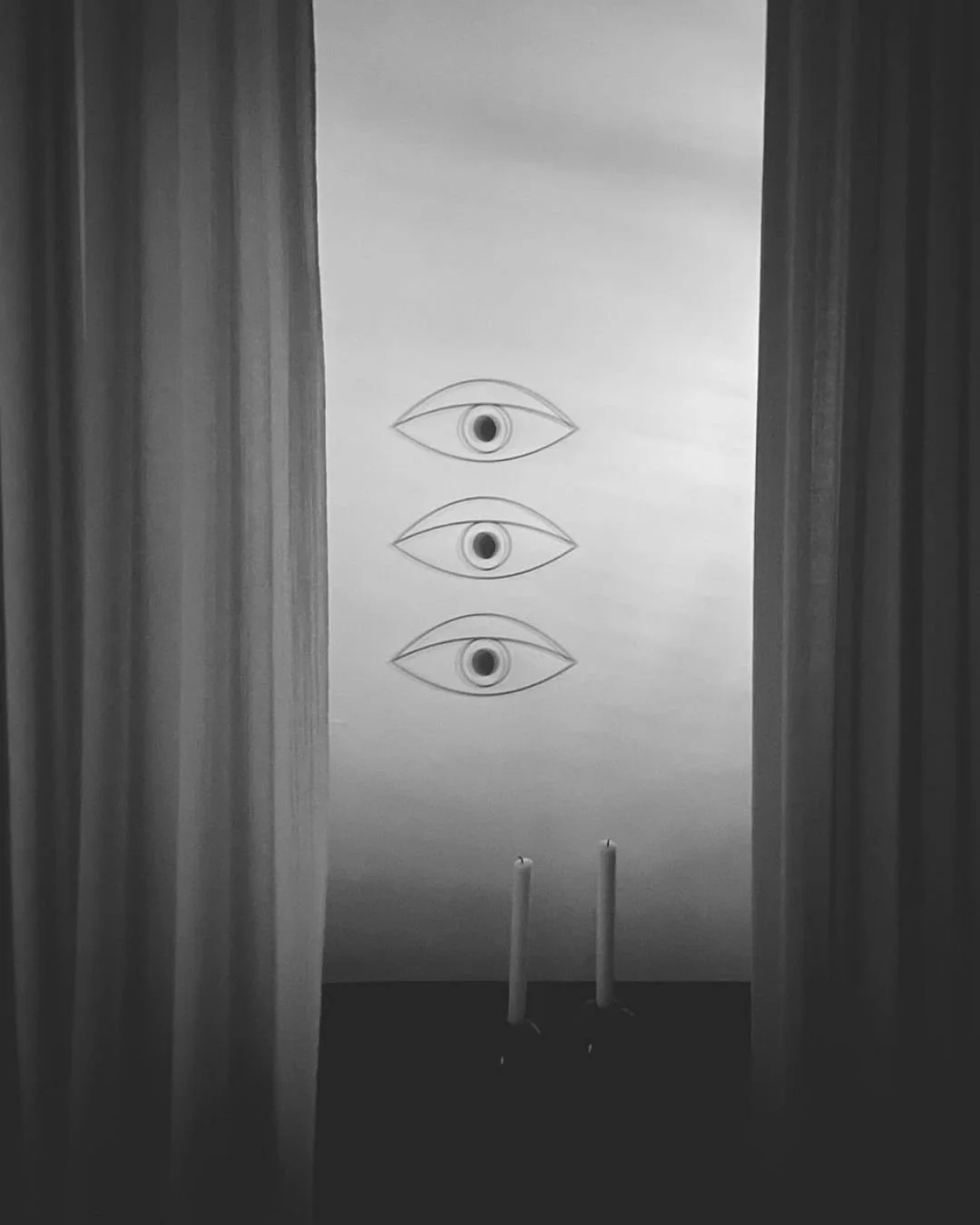 Schwarzes und weißes Bild mit drei Augen auf einer weißen Wand, zwei Kerzen stehen davor, die Szene ist durch Vorhänge eingerahmt.
