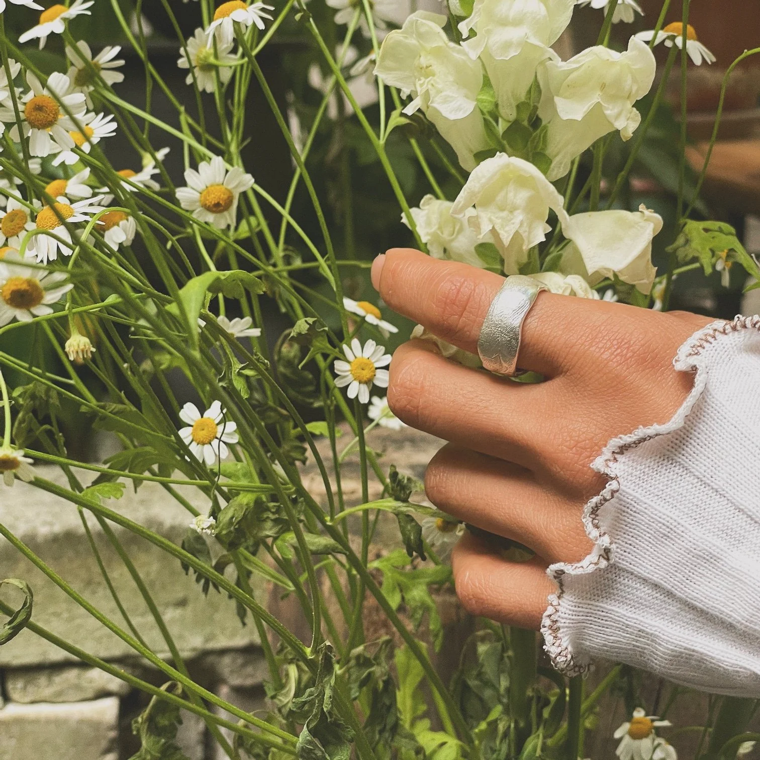 Person wearing a silver ring and a white, frilly sleeve touching white sweet pea flowers surrounded by small daisies with yellow centers.