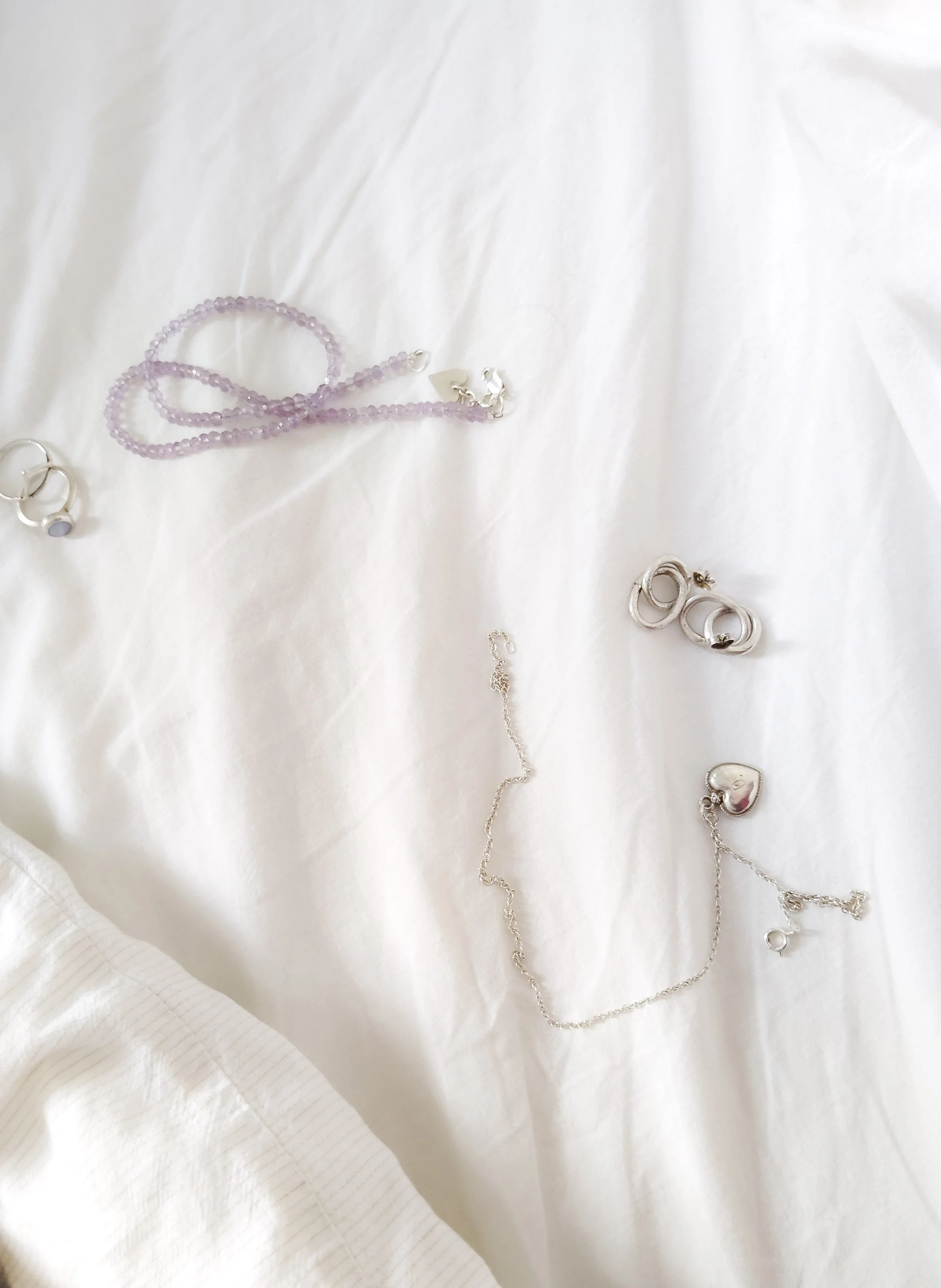 Collection of jewelry, including a purple beaded necklace and silver rings, placed on white fabric.