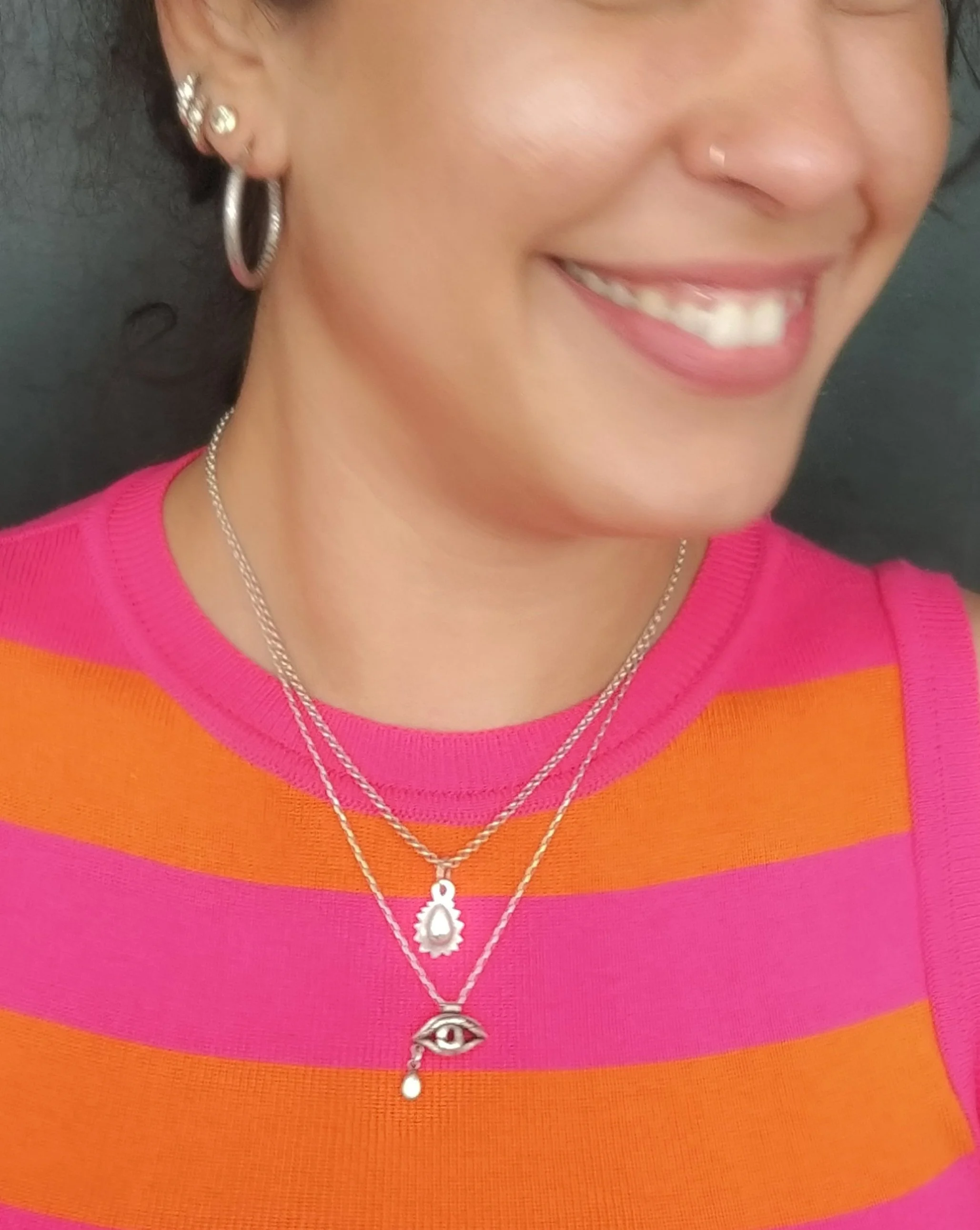 Close-up of a woman's smile showing her teeth, wearing a pink and orange striped top, with layered necklaces and earrings, and a small nose piercing.
