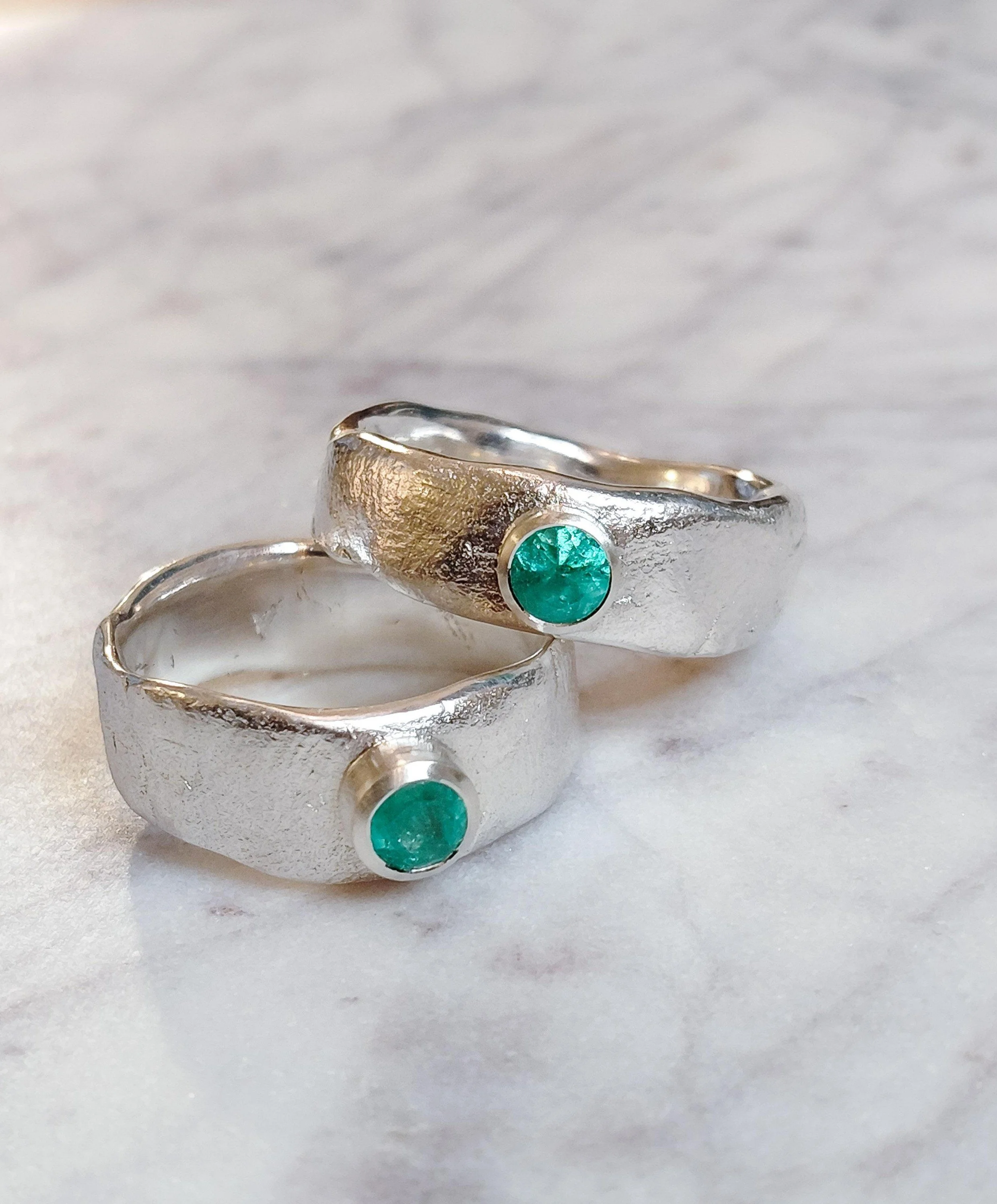 A silver ring with a green gemstone resting on a white surface, with a second silver ring partially visible beneath it.