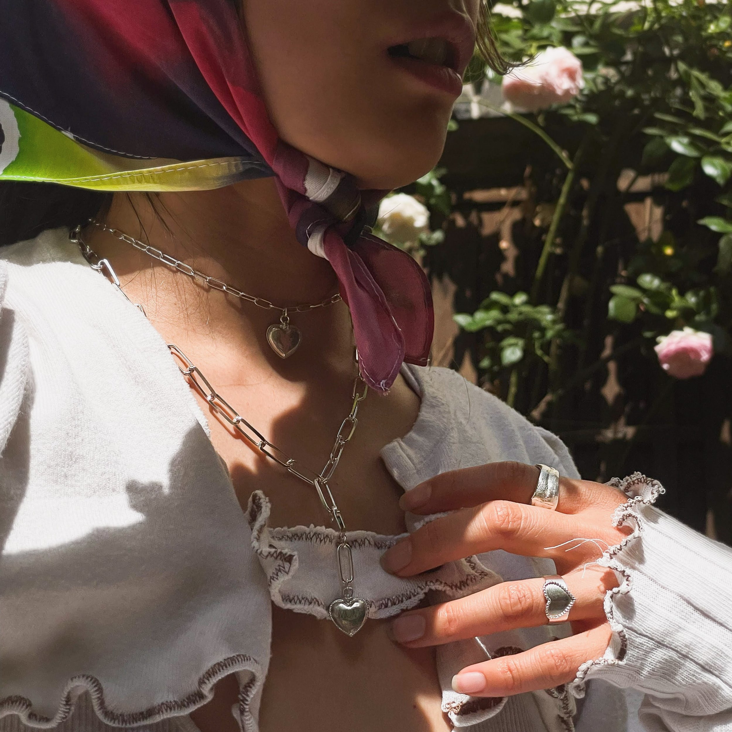 Close-up of a person's face, showing lips and part of the nose, wearing a white, embroidered blouse and layered jewelry including a chain with a heart pendant, rings, and a bracelet. The person is holding a lace scarf or cloth, with greenery and pink