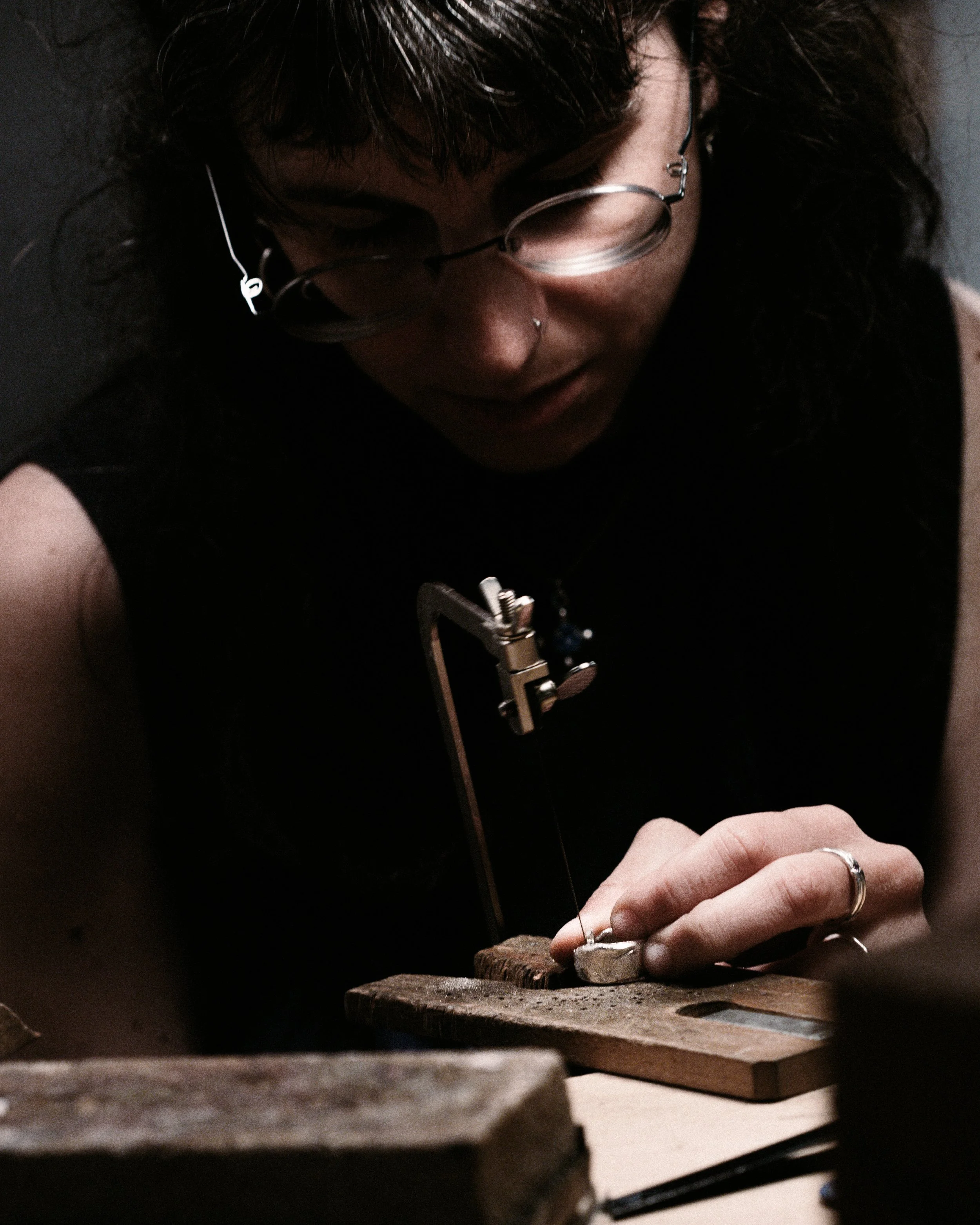 Close-up of a person with dark curly hair, wearing glasses, a nose piercing, and rings, working with a small handsaw on a silver unfinished ring, in a jewelry-maker's workshop.