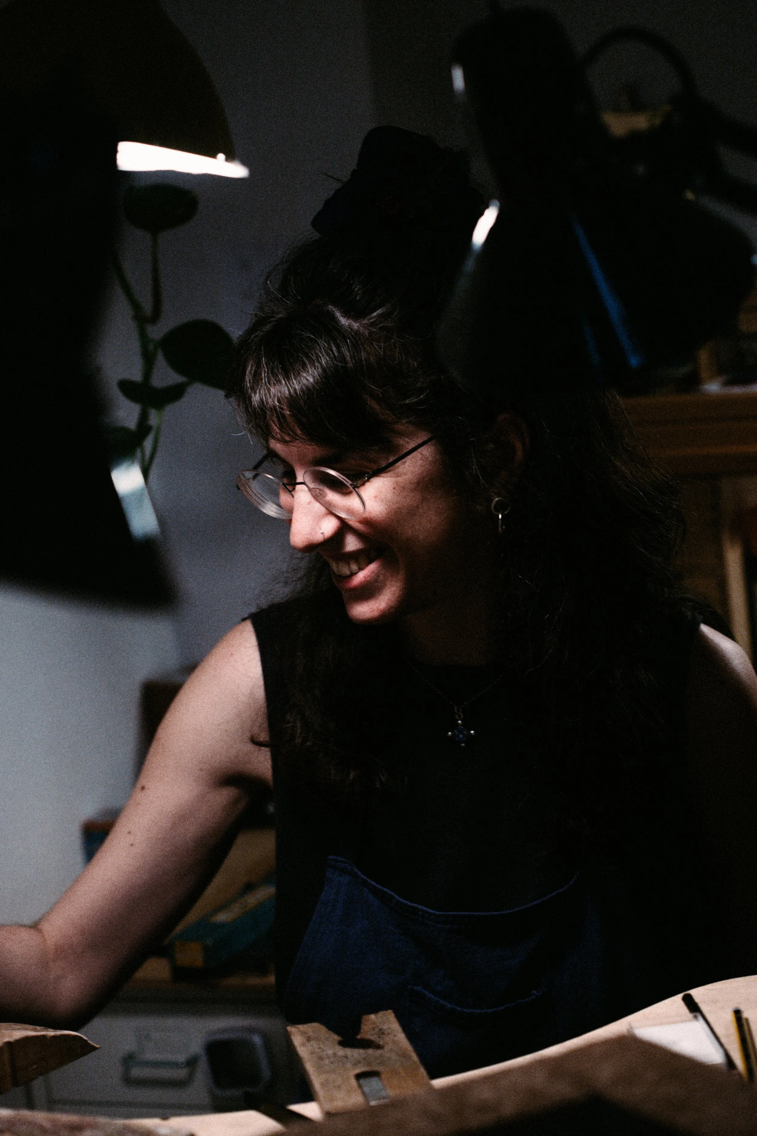 Woman with glasses smiling, working on a woodworking project in a dimly lit room.