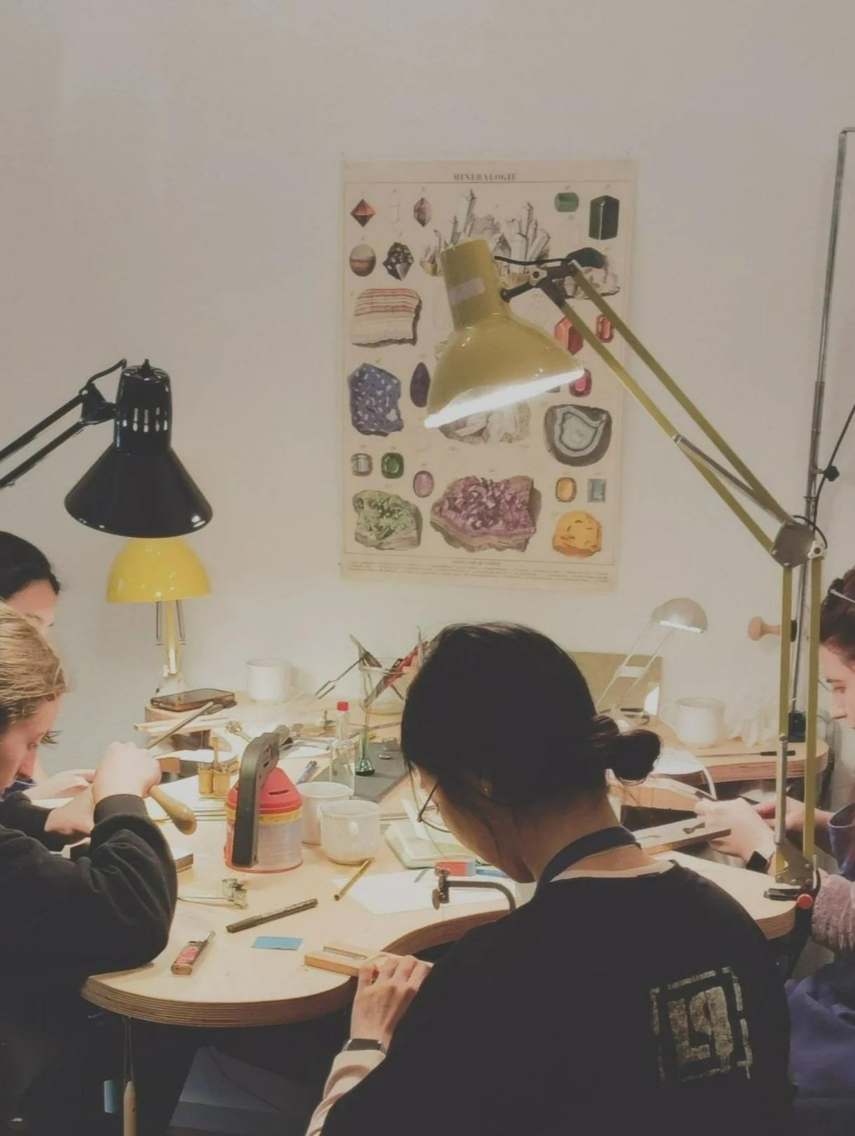 People working at a cluttered table with lamps, tools, and art materials, and a poster of minerals on the wall behind them.