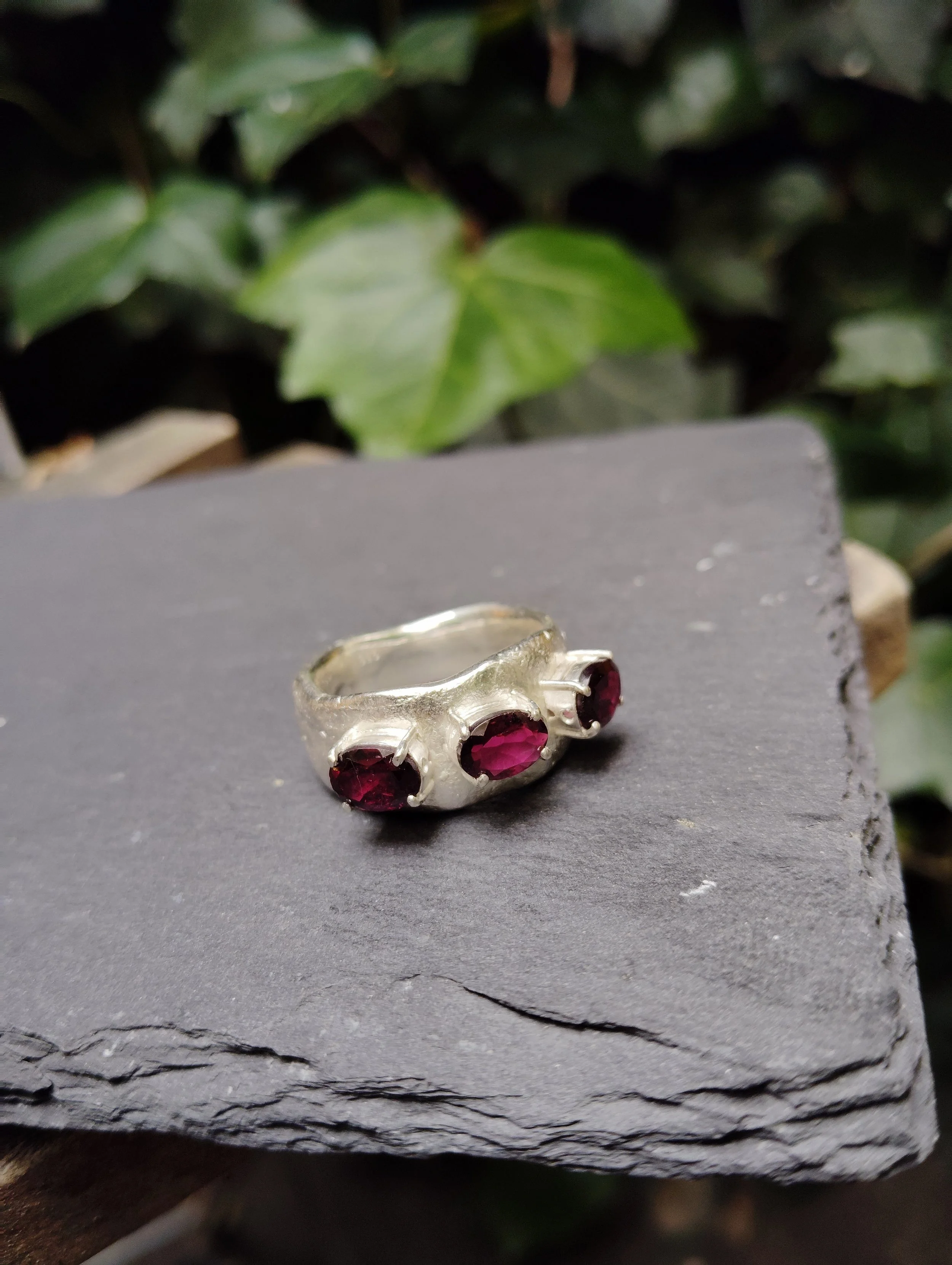 Silver ring with three red gemstones on a black surface, with green leaves in the background.