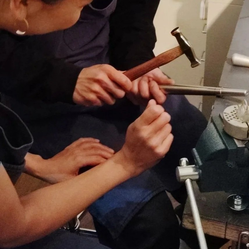 Two people working on their wedding rings with tools, including a hammer and a ring mandrel, at a workbench.