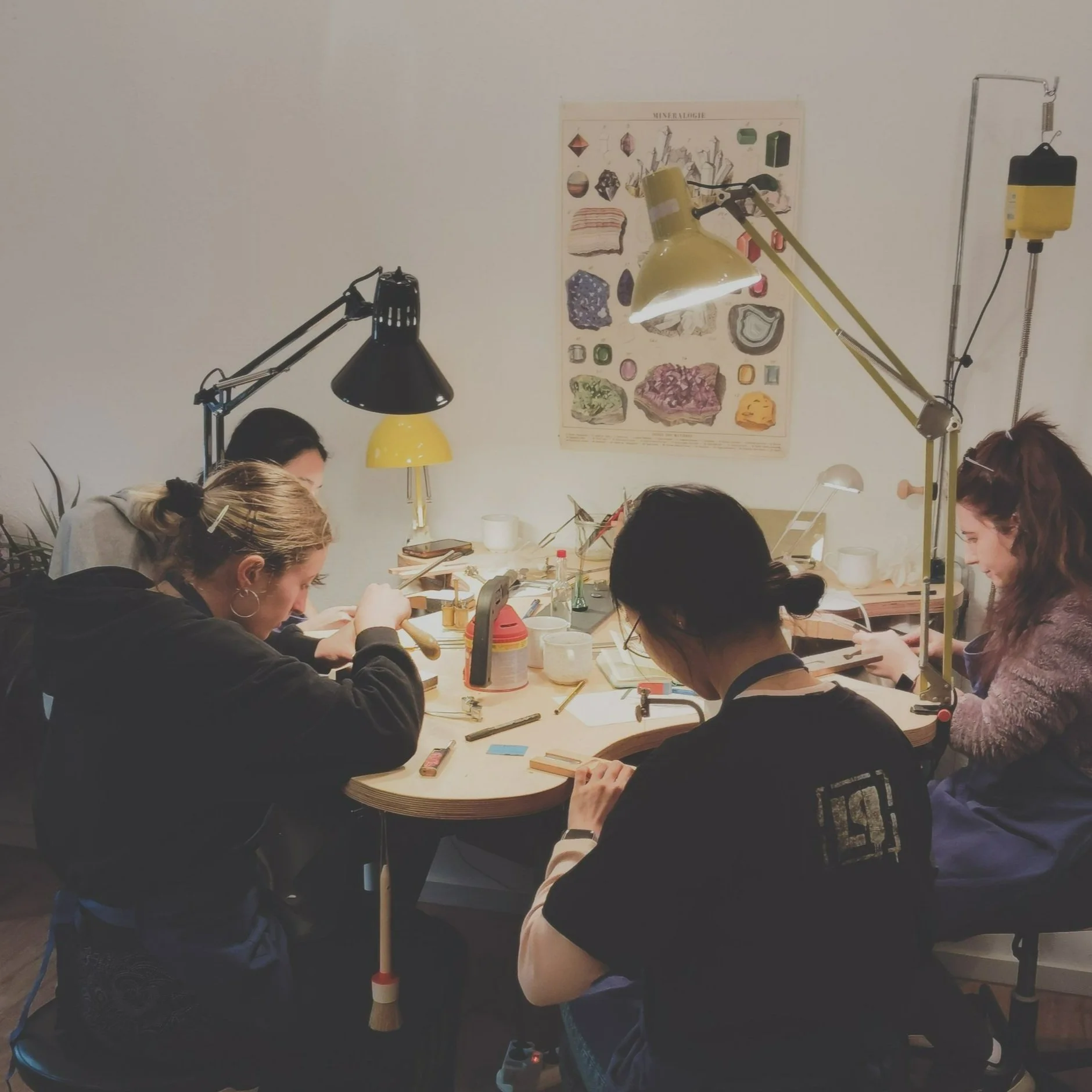 A group of people seated around a table working on theirjewelry projects, with yellow and black lamps overhead and a mineralogy poster on the wall in the background.