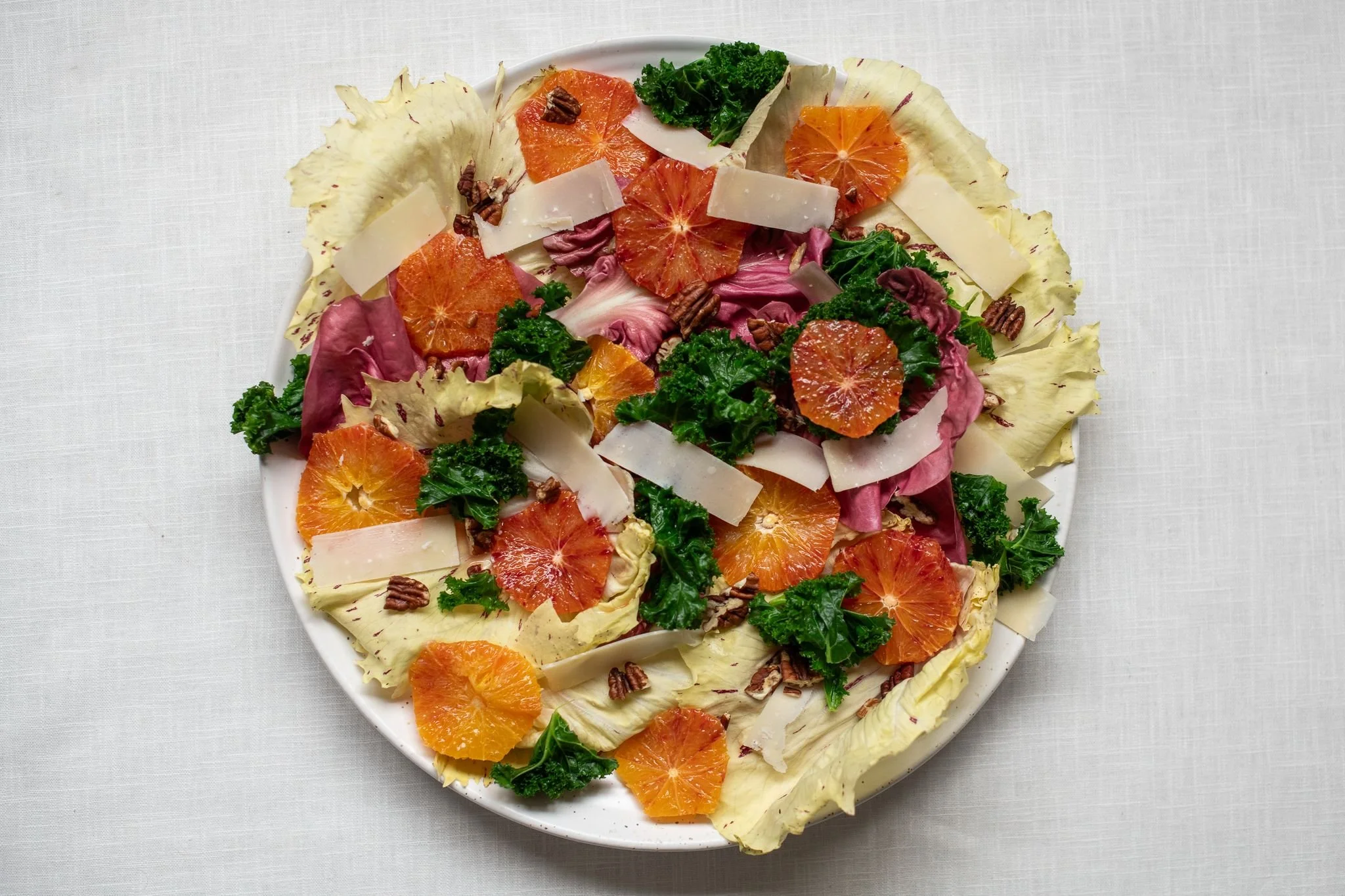 Salad with lettuce, mandarins, cheese, kale, pecans, and pink radicchio on a white plate.