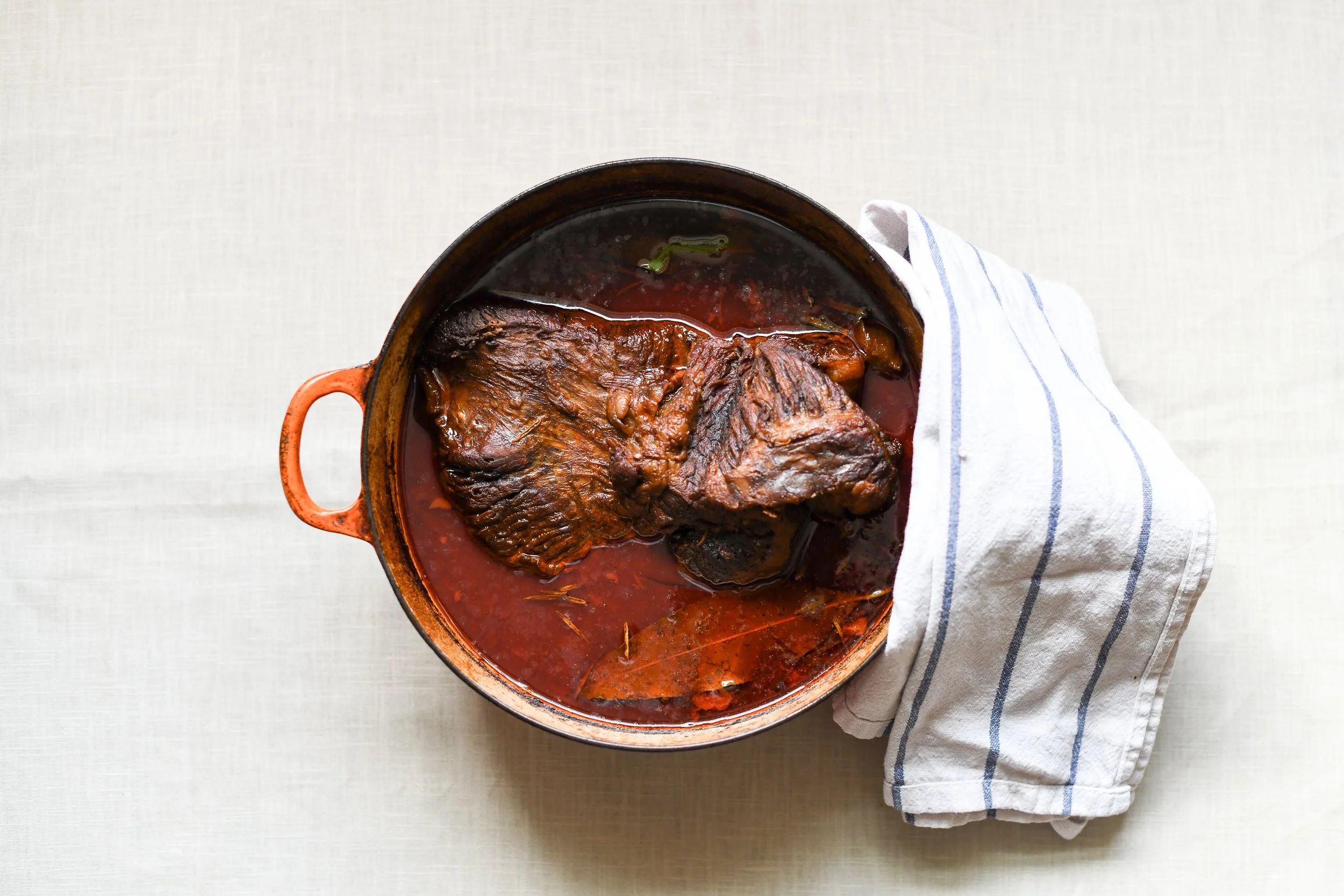 Pot of cooked beef stew with a large piece of meat, in a tomato-based broth, placed on a white surface with a striped cloth towel beside it.