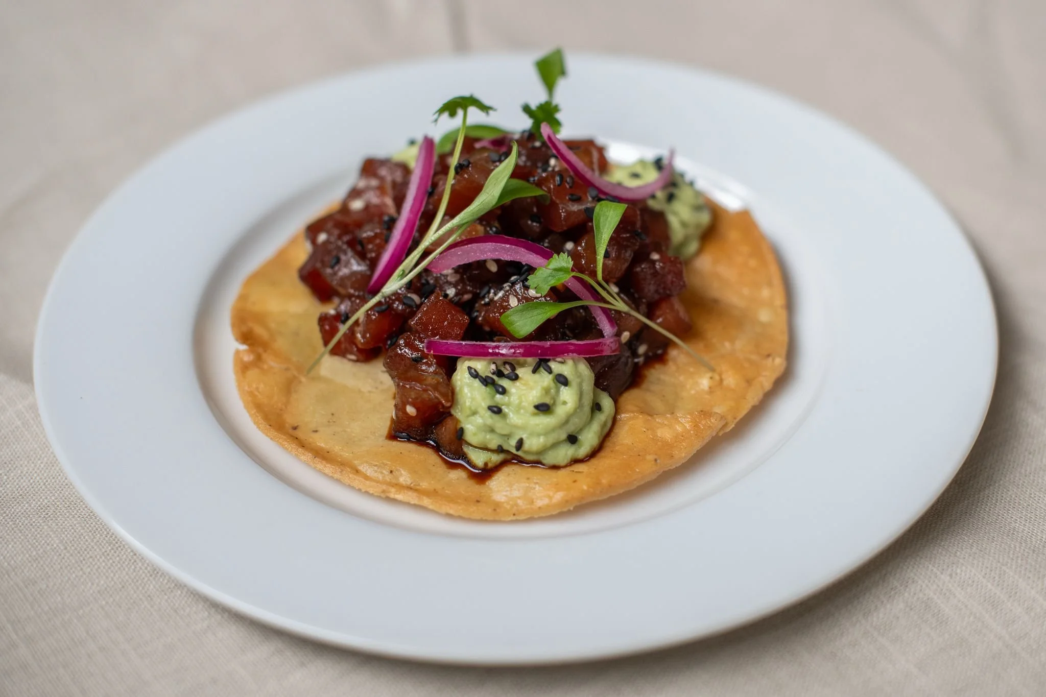 Tuna tartare served on a crispy tortilla chip, topped with sliced red onions, microgreens, black sesame seeds, and guacamole, on a white plate.