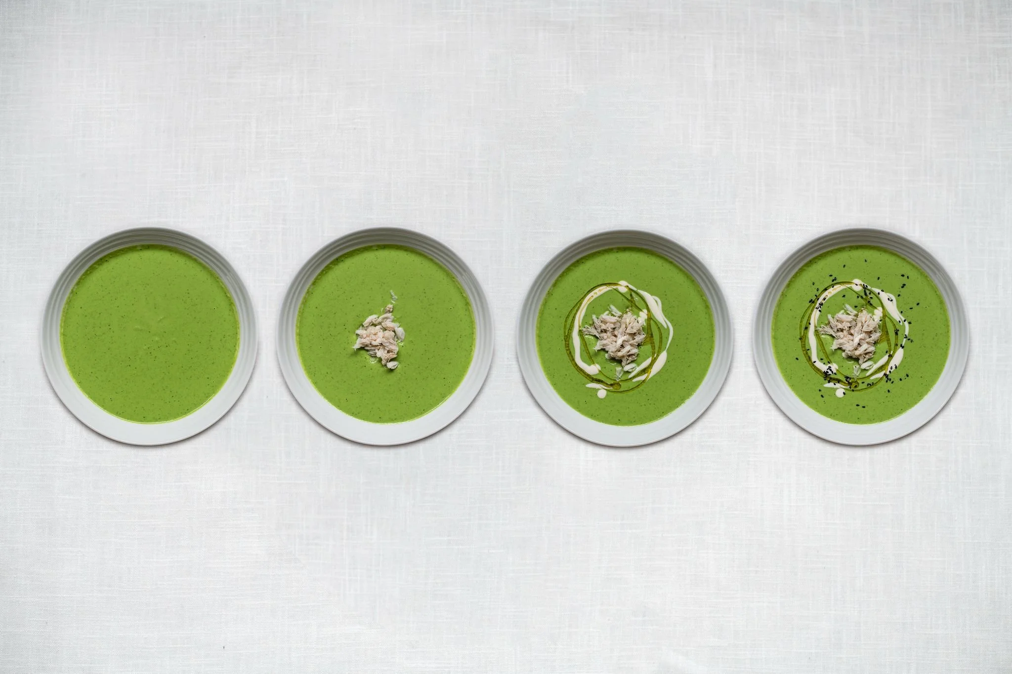 Four bowls of green soup arranged in a row on a white surface, garnished with shredded chicken, and drizzled with different sauces and spices.