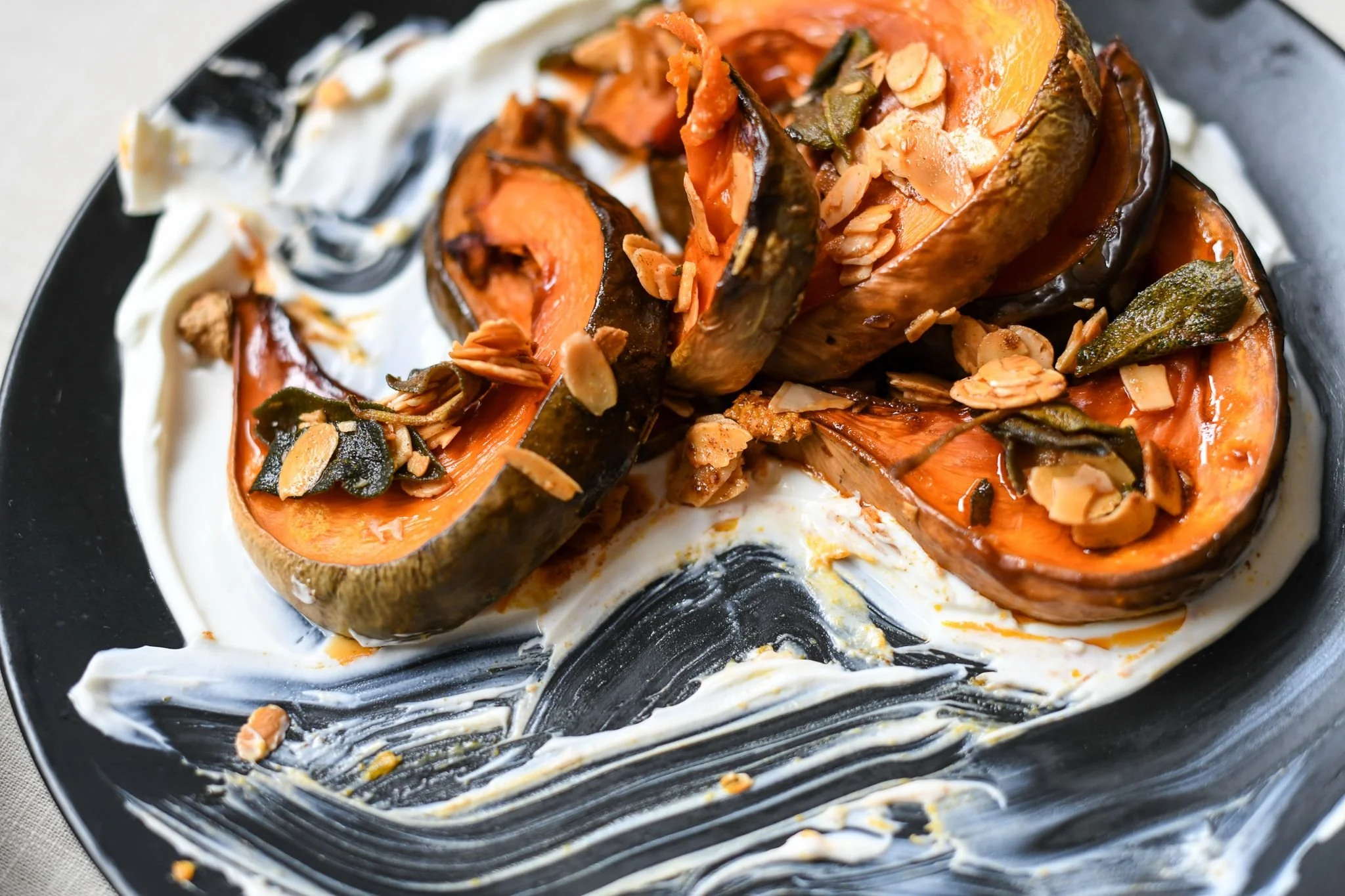 Halved butternut squash topped with sliced almonds and sage leaves, set on a black plate with swirls of cream and sauce.