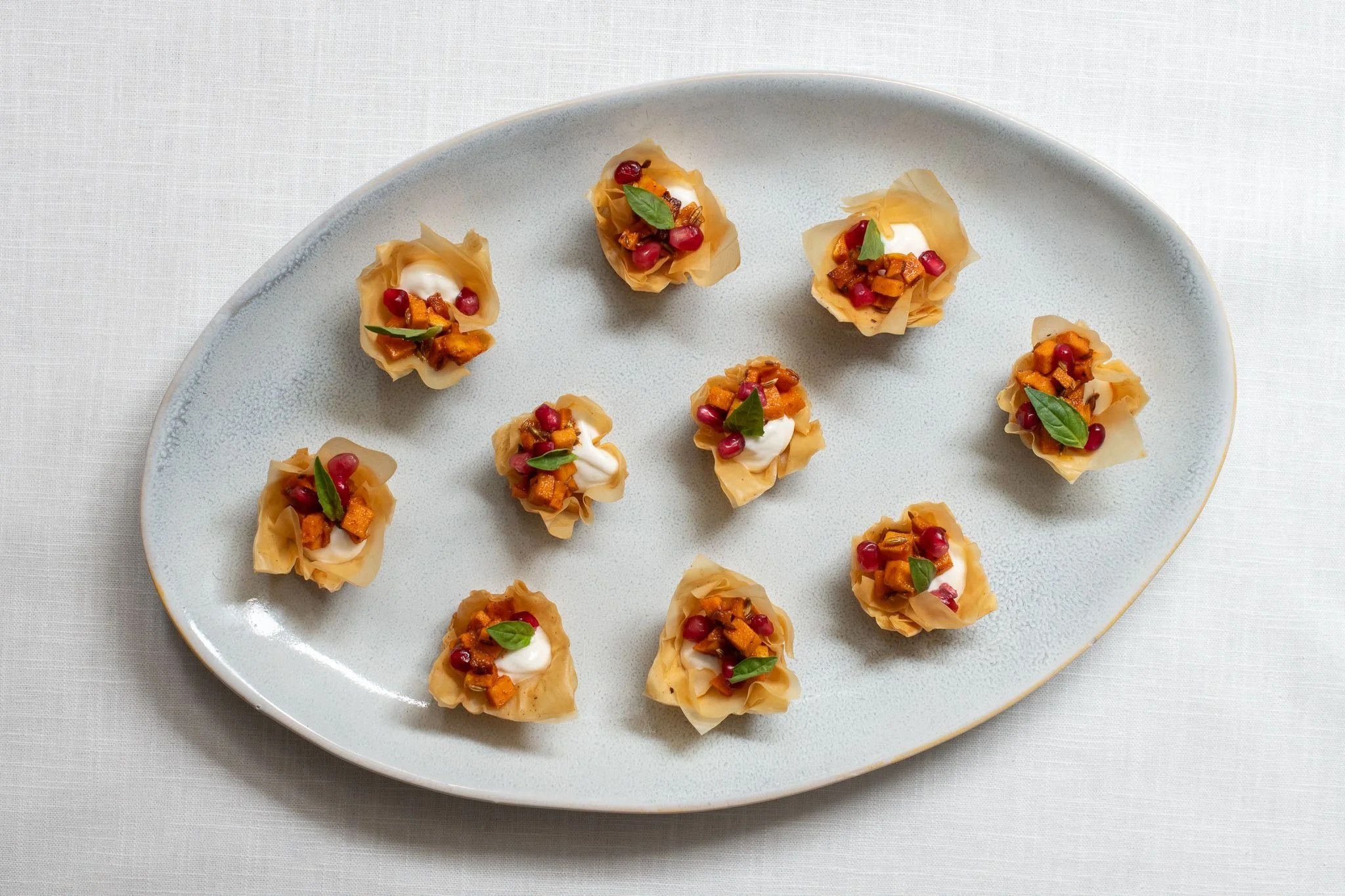 Appetizer served on a white oval plate with ten small, crispy pastry cups filled with roasted vegetables, garnished with dollops of white sauce and fresh green leaves.