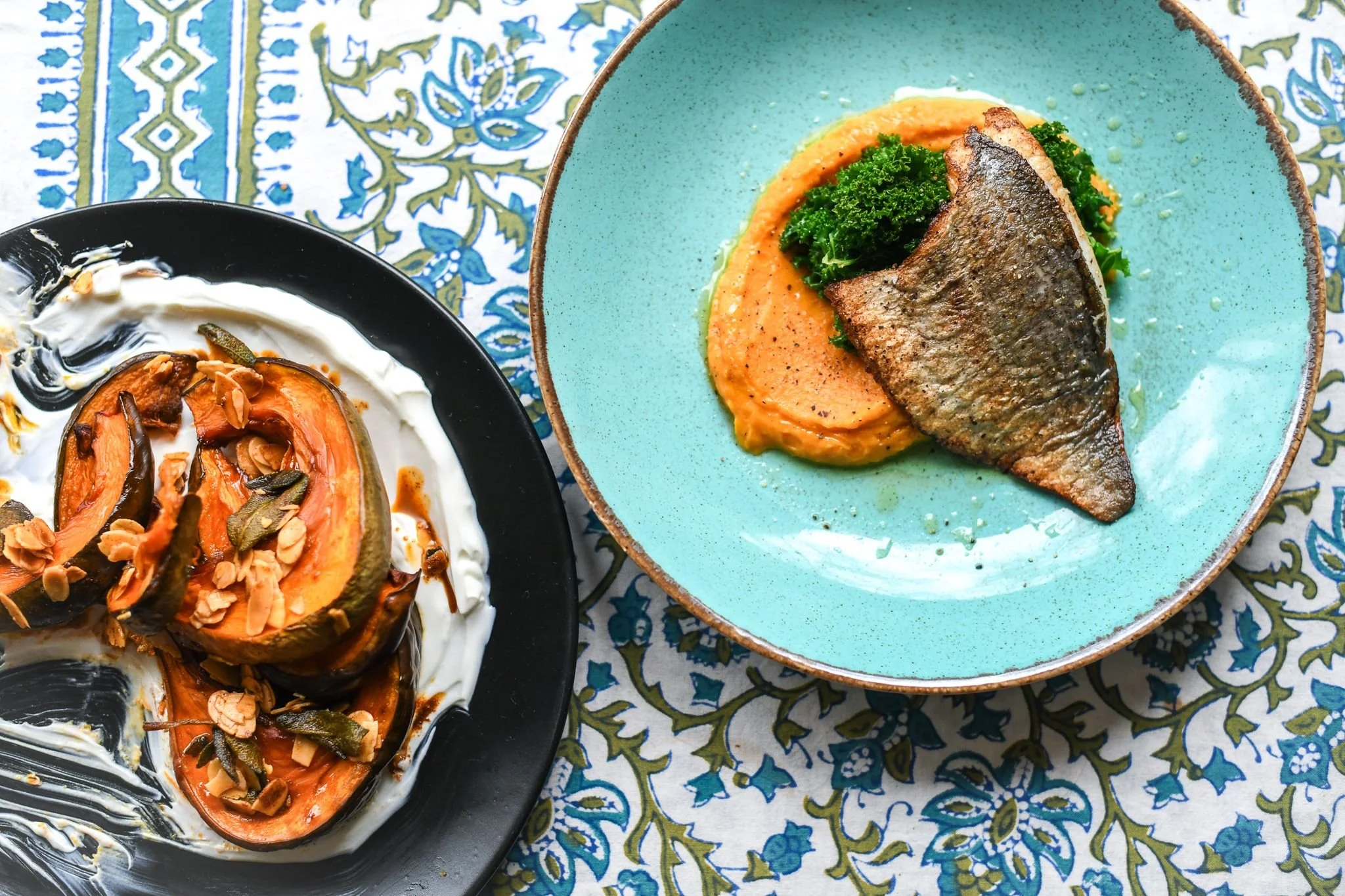 A plate with baked squash topped with slivered almonds and seeds on a black plate, and a blue ceramic plate with a fish fillet on a bed of mashed sweet potatoes and greens, on a patterned tablecloth.